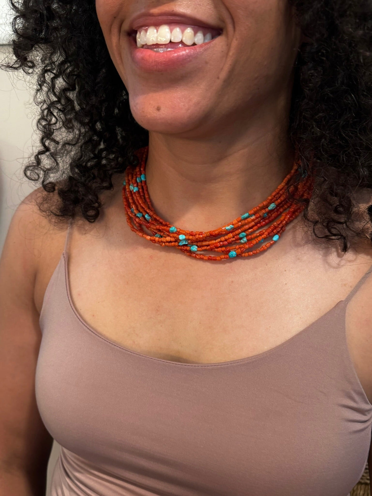 A close-up of a woman wearing a multi-strand beaded necklace with orange-red beads and small turquoise blue accents, paired with a taupe-brown camisole top.