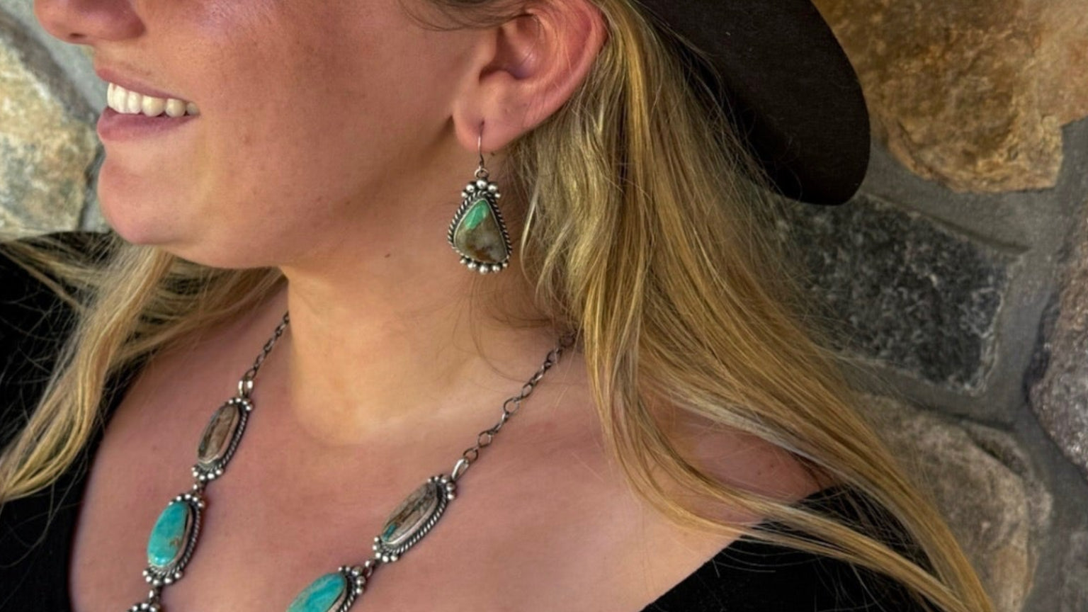 A woman wearing a black wide-brimmed cowboy hat, a black velvet top, and a turquoise and silver statement necklace, posing in front of a rustic stone wall.