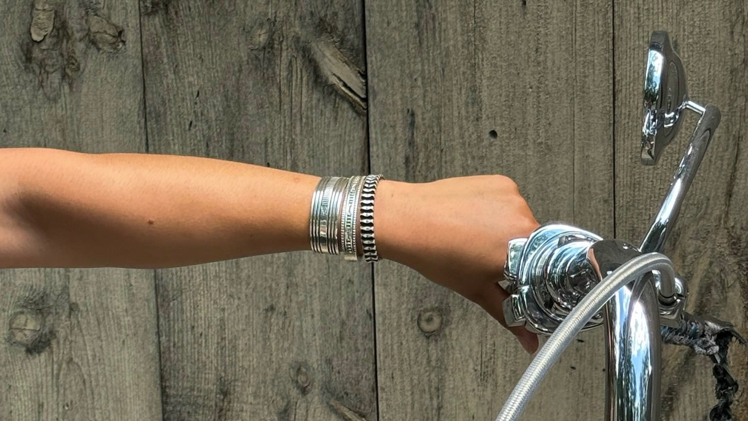 A close-up of a person's arm and hand wearing a silver-toned beaded bracelet and watch, touching the chrome handlebar of a motorcycle in front of a rustic wooden fence.