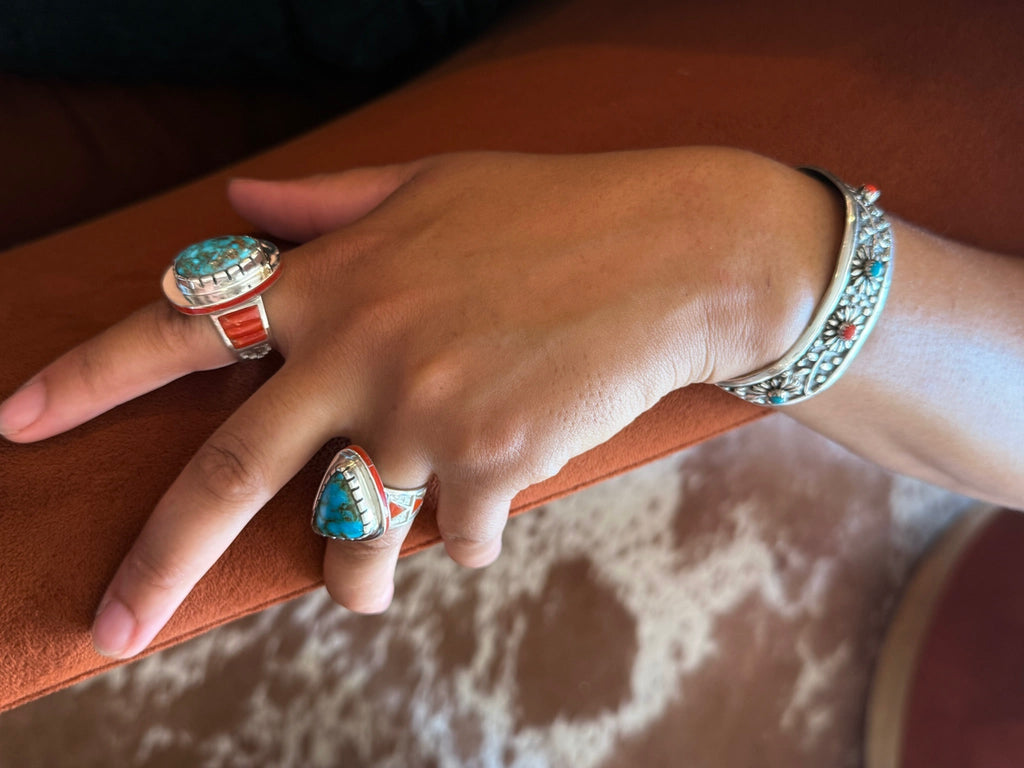 A close-up of a hand wearing three silver-toned rings and a matching cuff bracelet, each featuring turquoise and coral inlay details, resting on a brown leather armrest.