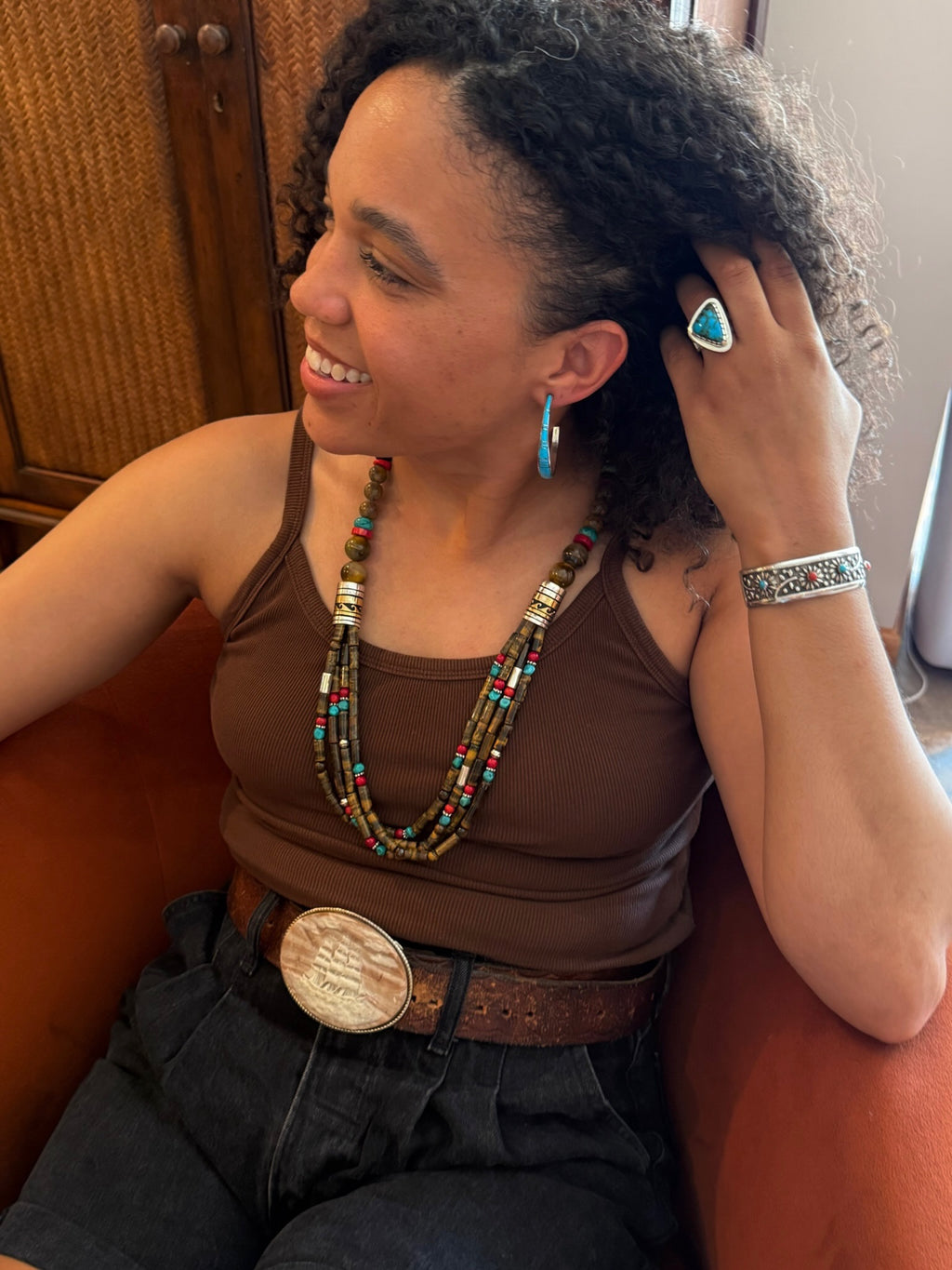 A woman with curly dark hair smiling while wearing a brown beaded necklace, turquoise hoop earring, silver ring, and a silver patterned bracelet. She is wearing a brown tank top, dark jeans with a large silver belt buckle, and a brown belt, sitting on an orange couch.