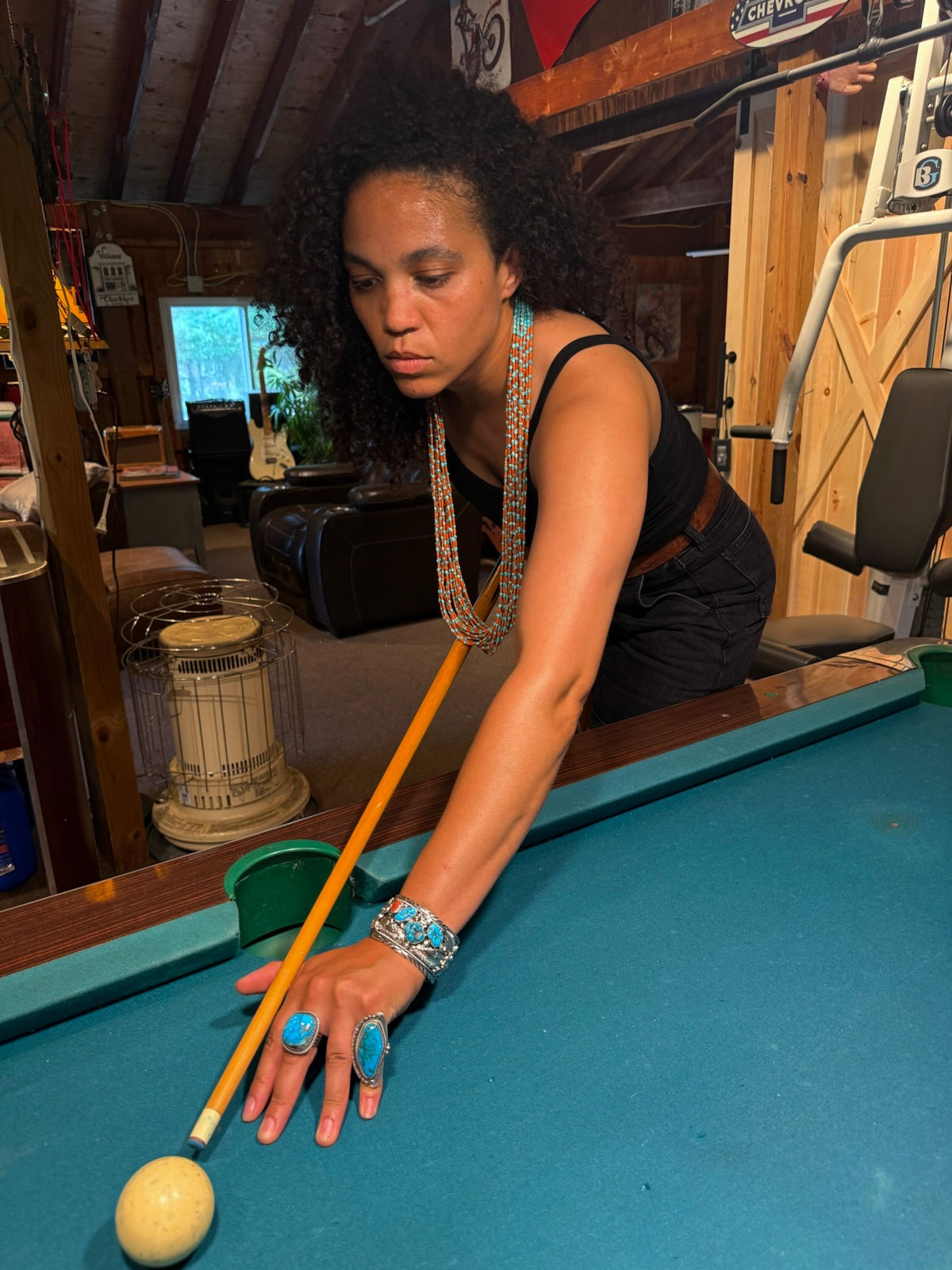 A woman with curly hair leans over a pool table, holding a yellow cue stick. She is wearing a black sleeveless top, a long silver beaded necklace, a turquoise bracelet, and turquoise rings.