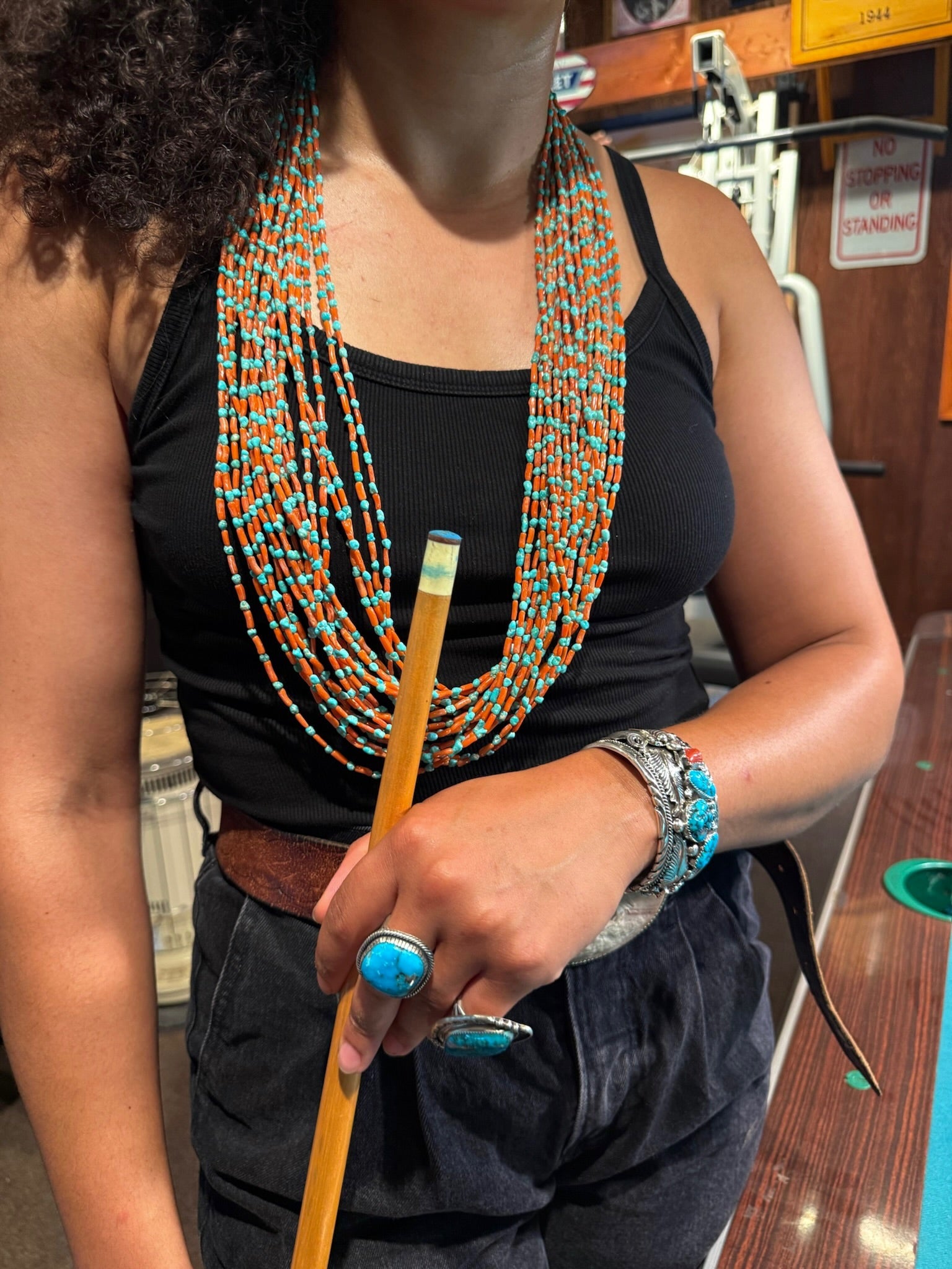 A person wearing a long strand of turquoise and orange beaded necklaces, silver bracelets, and turquoise rings, holding a yellow pencil in a classroom setting.