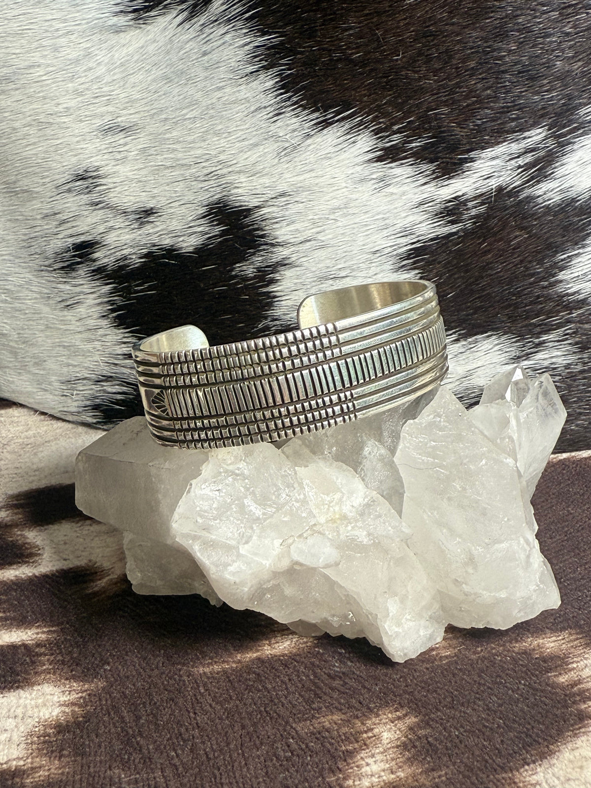 A wide silver cuff bracelet with textured horizontal grooves, displayed on a clear crystal cluster over a black and white cowhide surface.