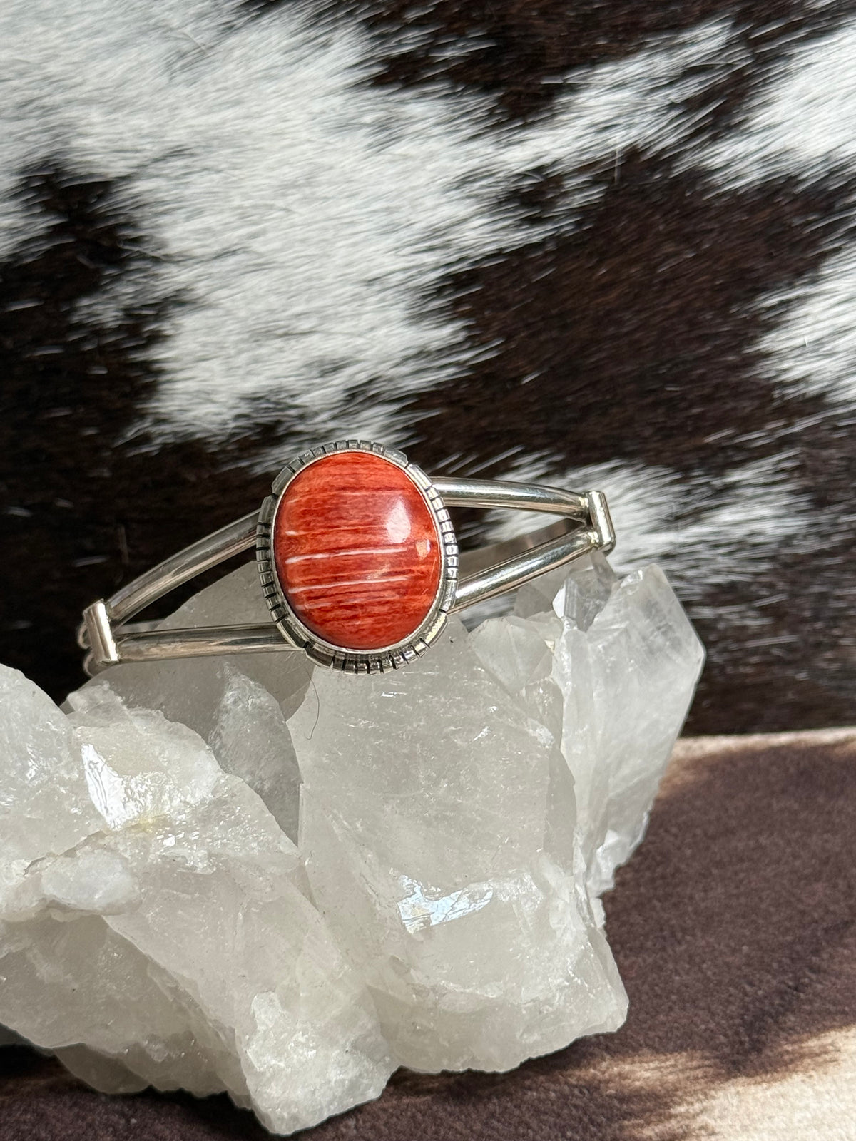 A silver-toned cuff bracelet with a large oval red-orange stone centerpiece, displayed on a clear crystal cluster against a black-and-white spotted fur background.