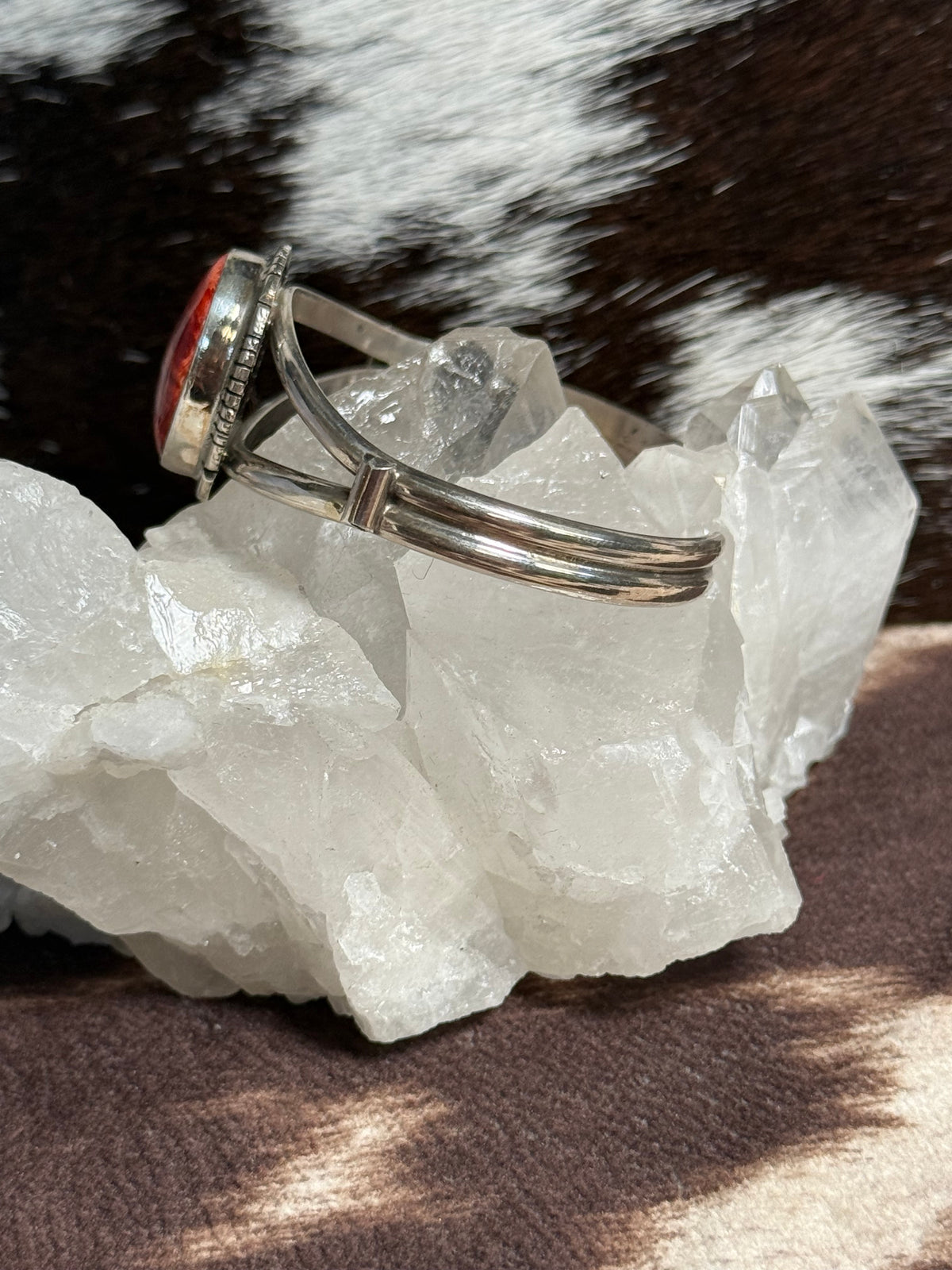 A silver-toned cuff with a red oval stone, displayed on a clear crystal cluster against a brown and white cowhide background.