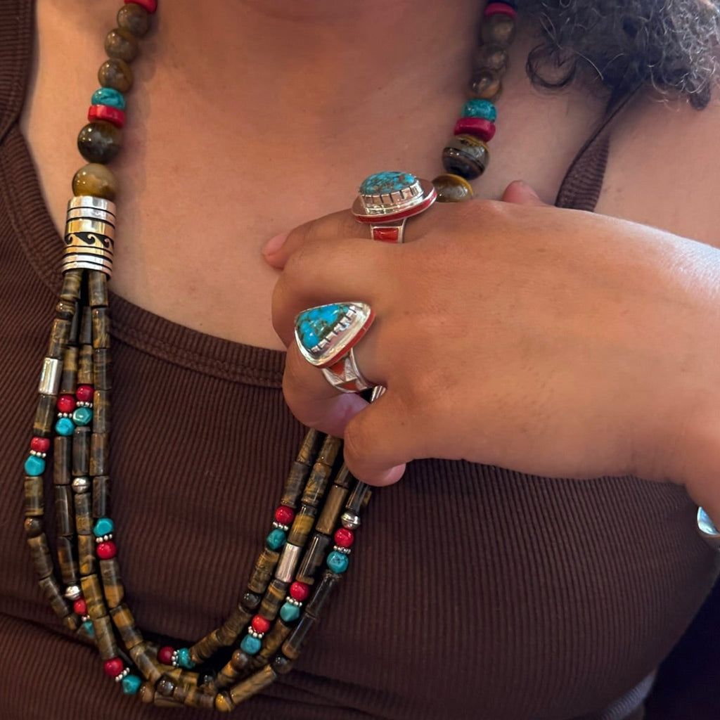 A close-up lifestyle shot of a person wearing a multi-strand beaded necklace with turquoise, red, and brown beads, along with silver rings.