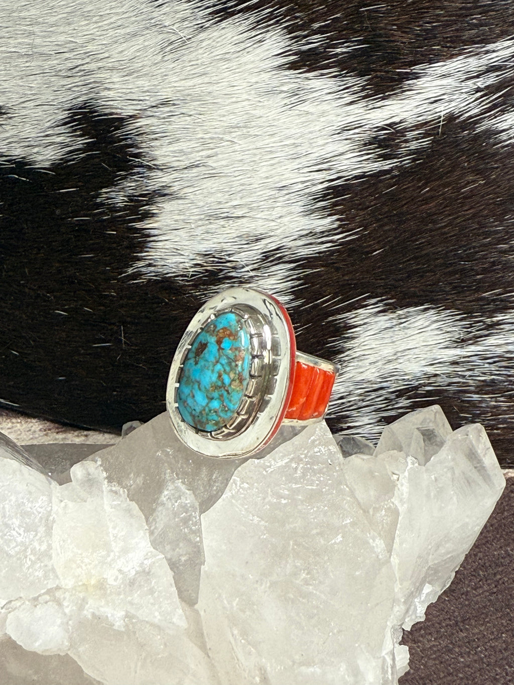 A turquoise and silver ring with a bright orange-red band, displayed on clear quartz crystals against a black and white cowhide background.