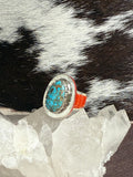 A turquoise and silver ring with a bright orange-red band, displayed on clear quartz crystals against a black and white cowhide background.