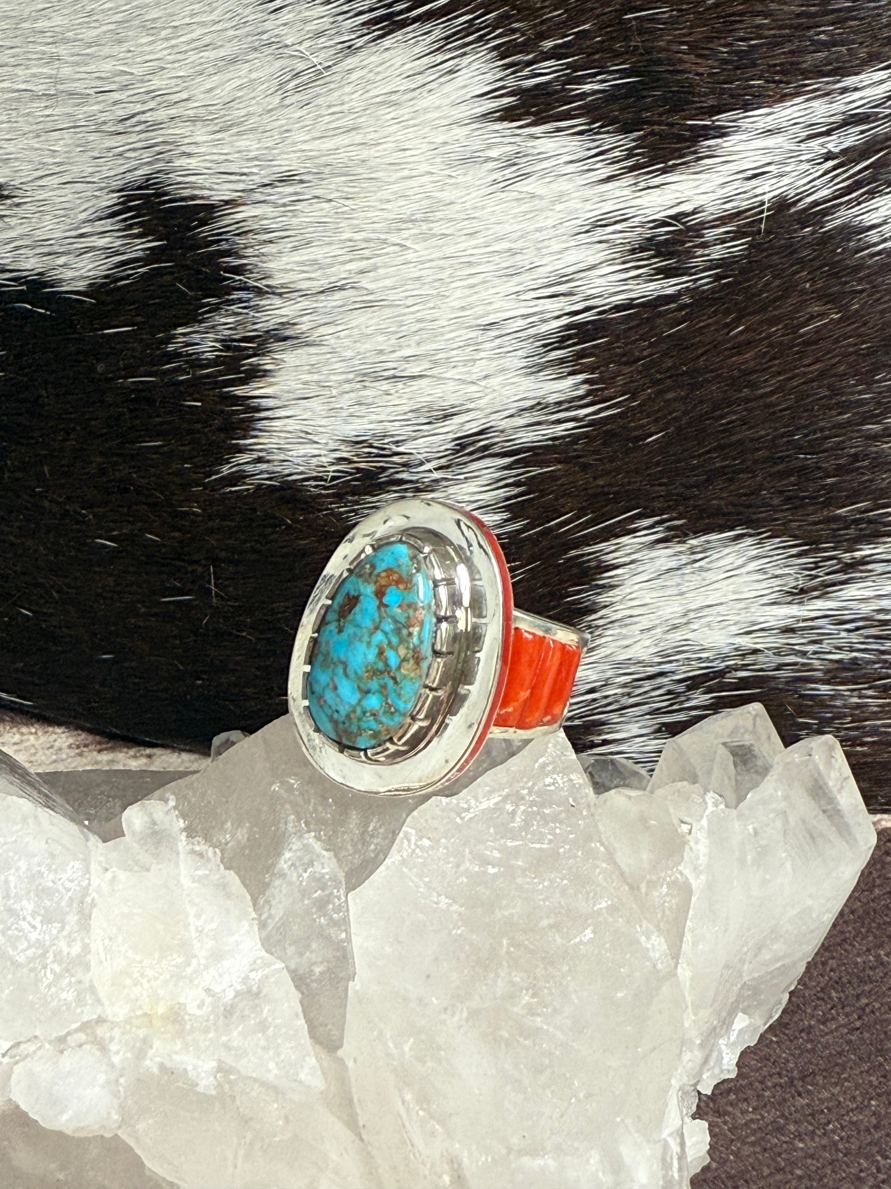 A turquoise and silver ring with a bright orange-red band, displayed on clear quartz crystals against a black and white cowhide background.