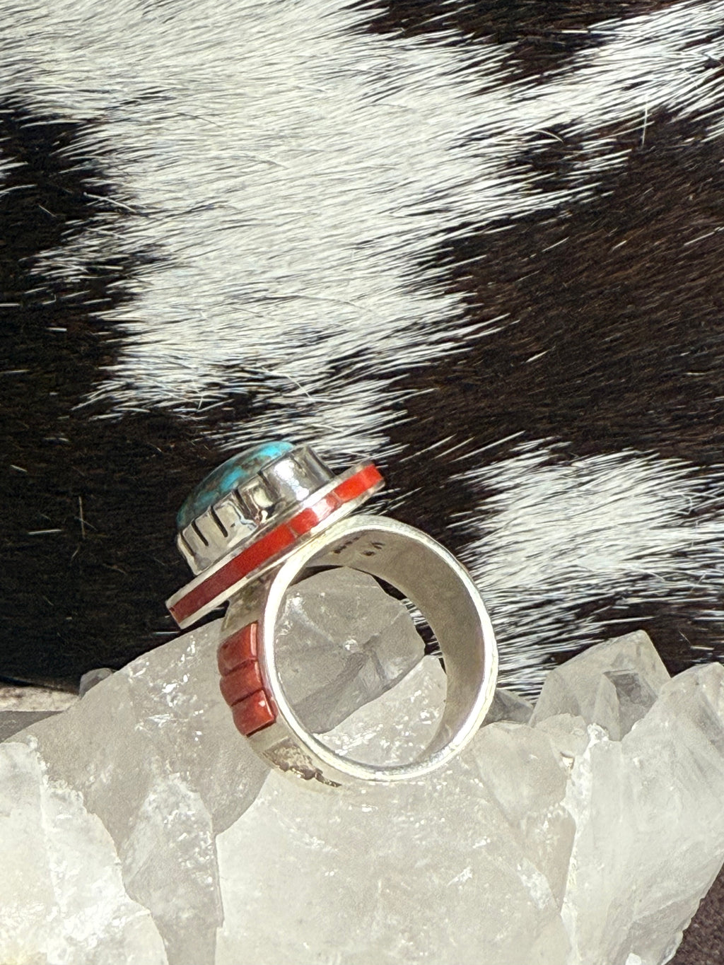 A silver-toned ring with a stacked geometric design, featuring turquoise, red, and white inlay details, displayed on clear quartz crystals against a black and white cowhide background.