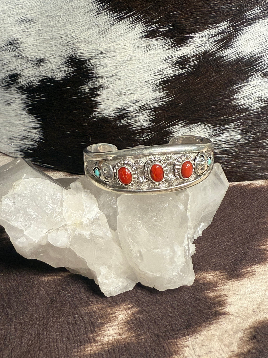 A silver cuff bracelet with red coral and turquoise stones, displayed on a white crystal cluster with a black and white cowhide background.