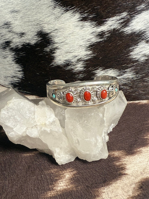 A silver cuff bracelet with red coral and turquoise stones, displayed on a white crystal cluster with a black and white cowhide background.