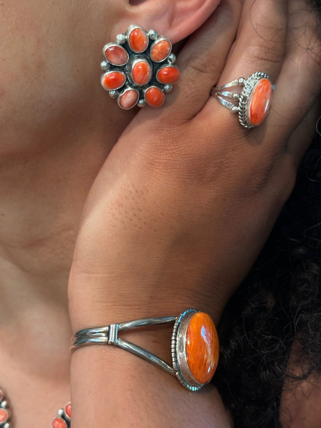 Close-up of a person wearing silver jewelry with vibrant orange stones, including a ring on the finger, a matching bracelet on the wrist, and a flower-shaped stud earring.