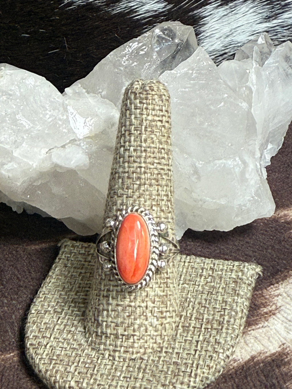 A silver ring with a large oval coral cabochon stone, displayed on a woven burlap ring holder. The holder is set against a background of white crystal clusters and dark brown fur.