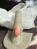 A silver ring with a large oval coral cabochon stone, displayed on a woven burlap ring holder. The holder is set against a background of white crystal clusters and dark brown fur.