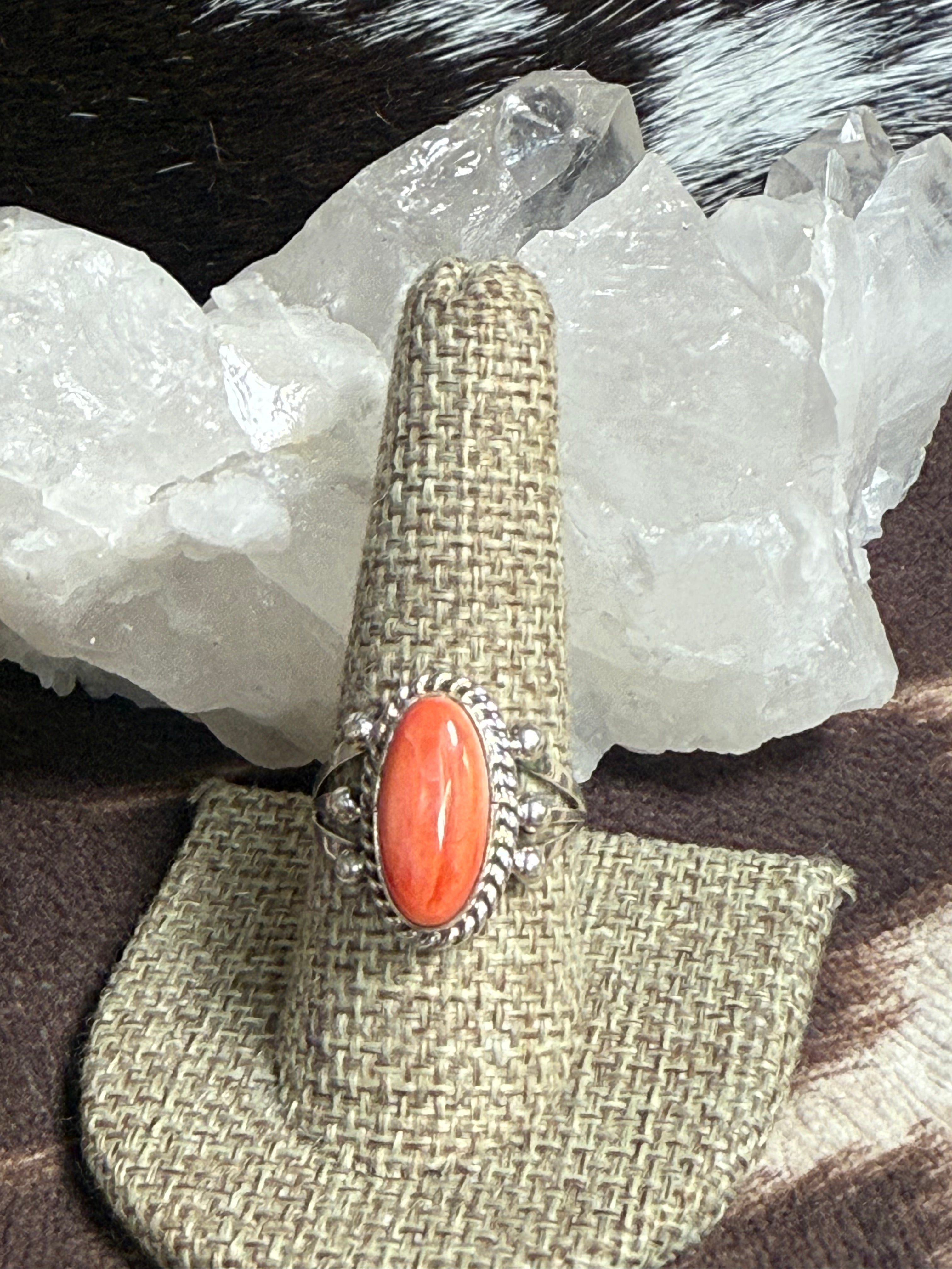 A silver ring with a large oval coral cabochon stone, displayed on a woven burlap ring holder. The holder is set against a background of white crystal clusters and dark brown fur.