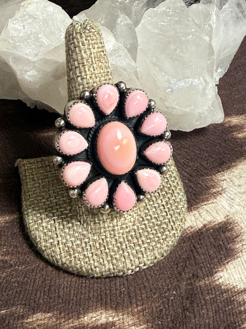A bohemian-style ring displayed on a woven burlap ring holder. The ring features a central oval pink cabochon surrounded by a circular cluster of smaller pink teardrop-shaped stones and silver-toned metal beads.