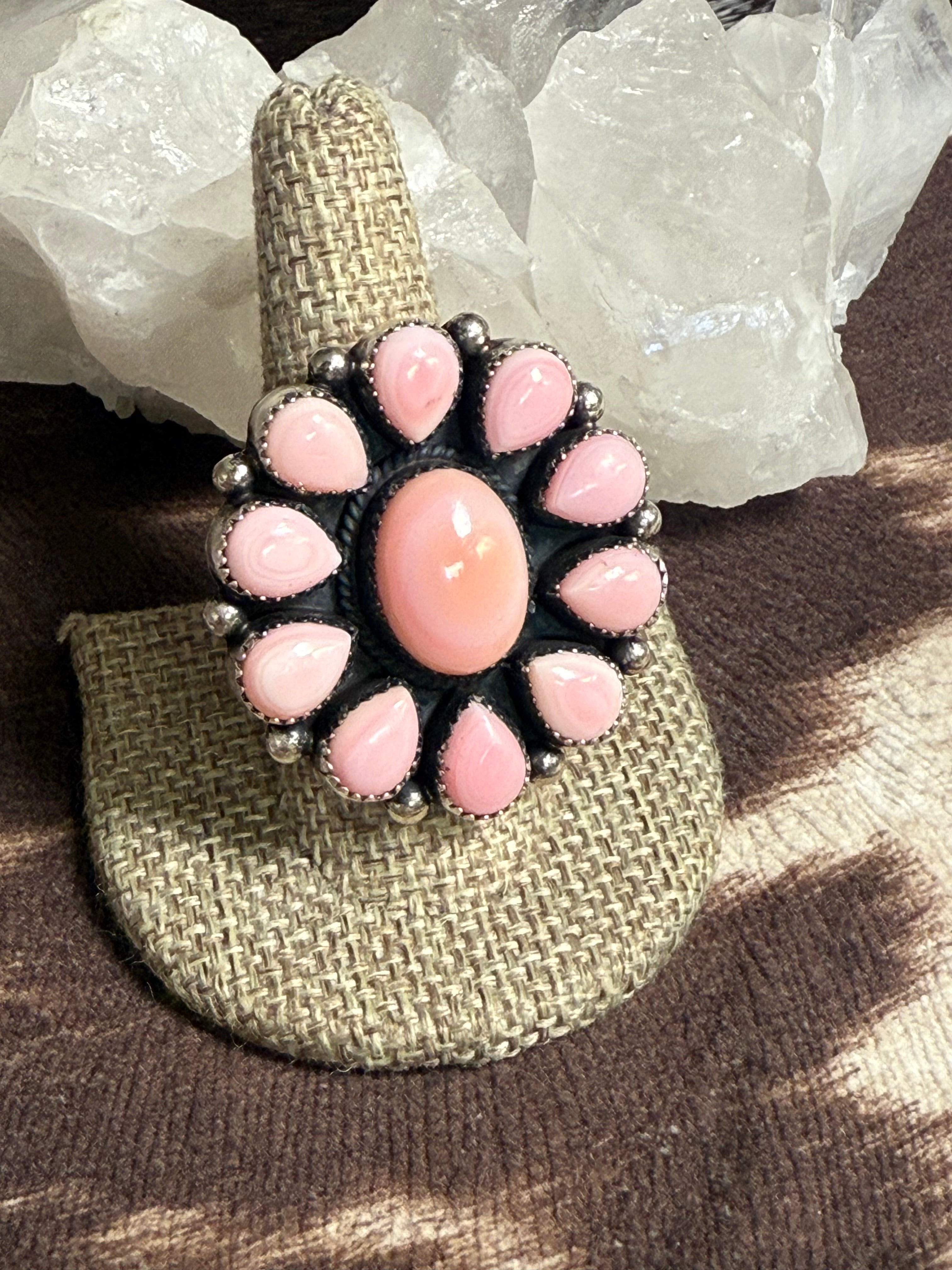 A bohemian-style ring displayed on a woven burlap ring holder. The ring features a central oval pink cabochon surrounded by a circular cluster of smaller pink teardrop-shaped stones and silver-toned metal beads.