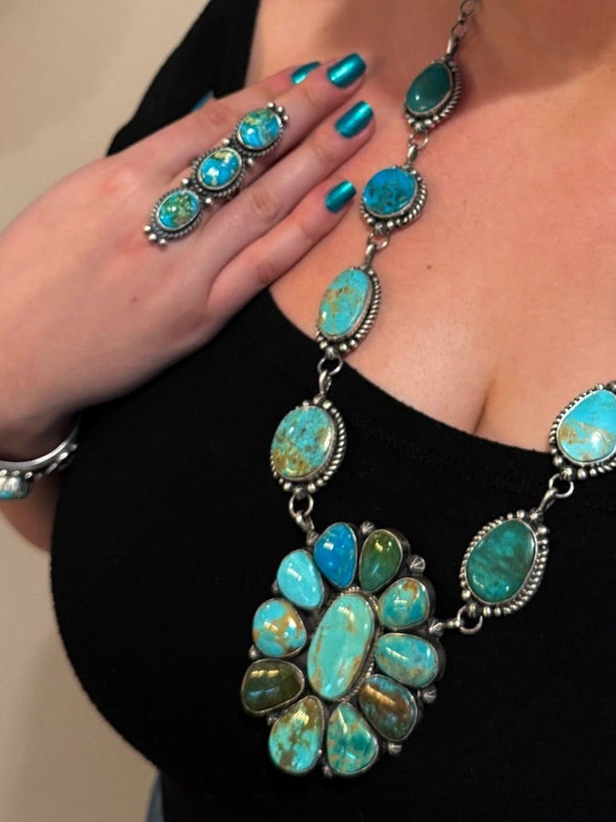A woman wearing a turquoise and silver jewelry set, including a necklace, earrings, and ring, paired with a black top and blue jeans in an indoor setting.
