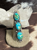 A silver-toned ring with three oval turquoise stones, displayed on a burlap ring holder in front of a clear crystal cluster.