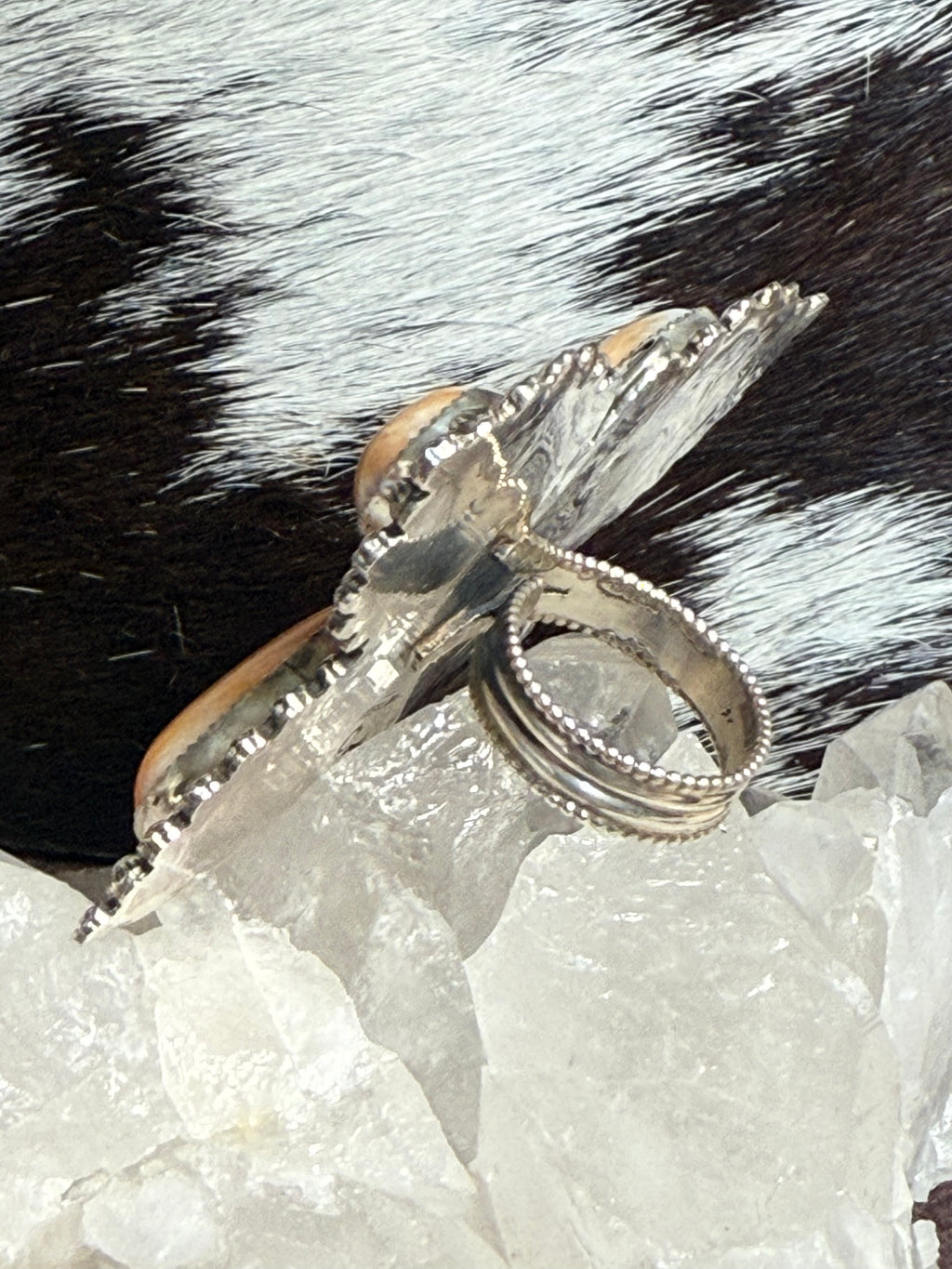 A silver-toned ring with a large butterfly-shaped setting and a small peach-colored stone, displayed on clear crystal against a black-and-white cowhide background.