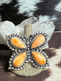 A butterfly-shaped silver-toned ring with four orange cabochon stones, displayed on a burlap-covered stand with clear quartz crystals behind it.