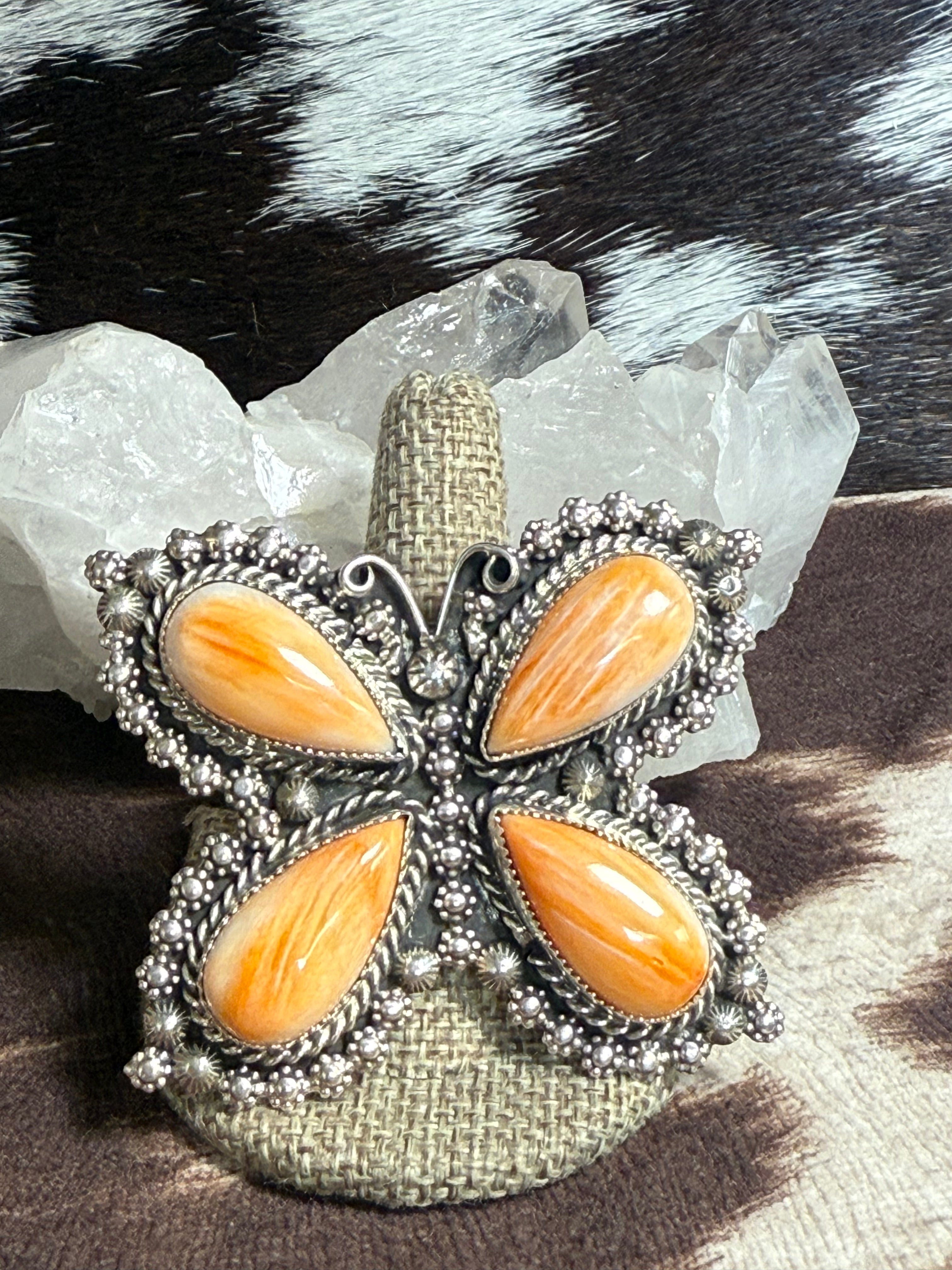 A butterfly-shaped silver-toned ring with four orange cabochon stones, displayed on a burlap-covered stand with clear quartz crystals behind it.
