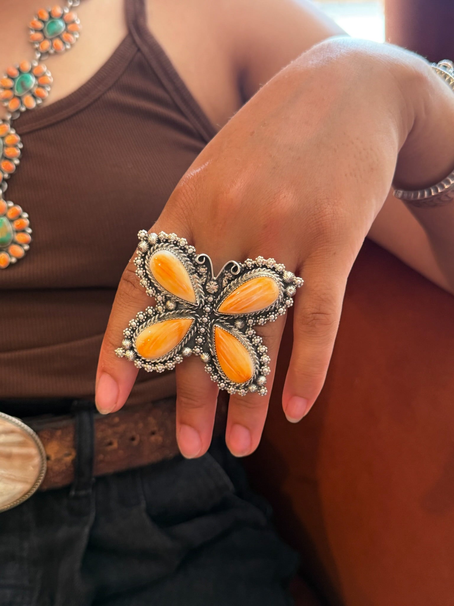 A close-up of a hand wearing a large butterfly-shaped ring with orange inlay and silver-toned beaded borders, worn over a brown tank top.