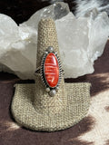 A silver ring with a carved red jasper cabochon, displayed on a burlap ring holder in front of white crystal clusters.