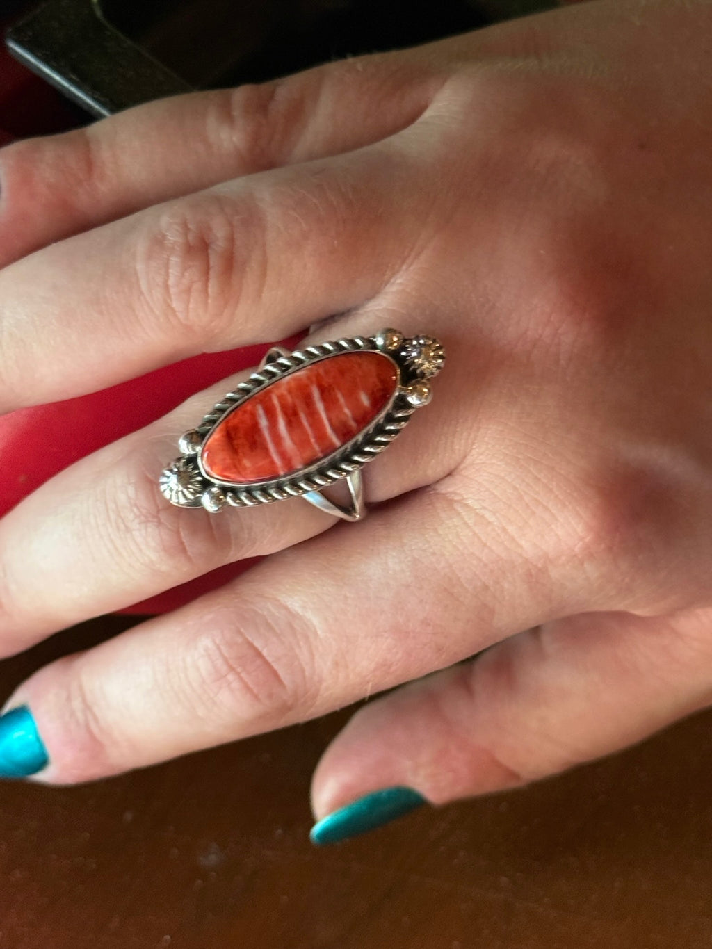 A close-up of a hand wearing a silver-toned ring with a large oval red-orange stone, photographed against a dark brown and red background.