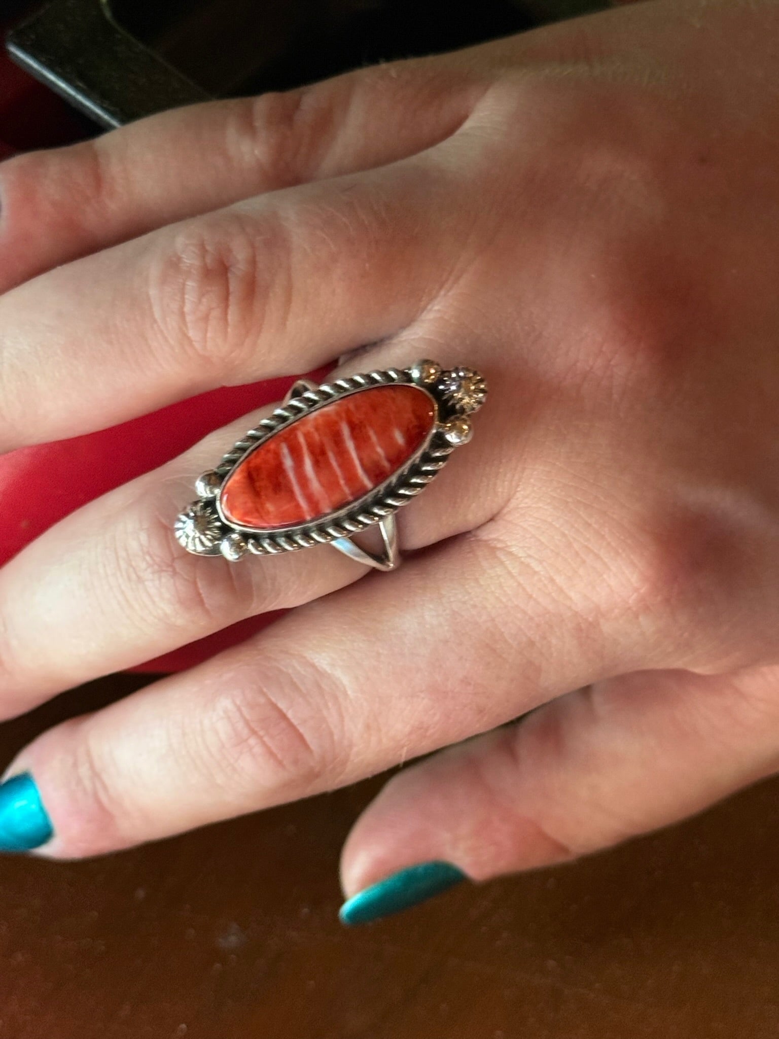 A close-up of a hand wearing a silver-toned ring with a large oval red-orange stone, photographed against a dark brown and red background.