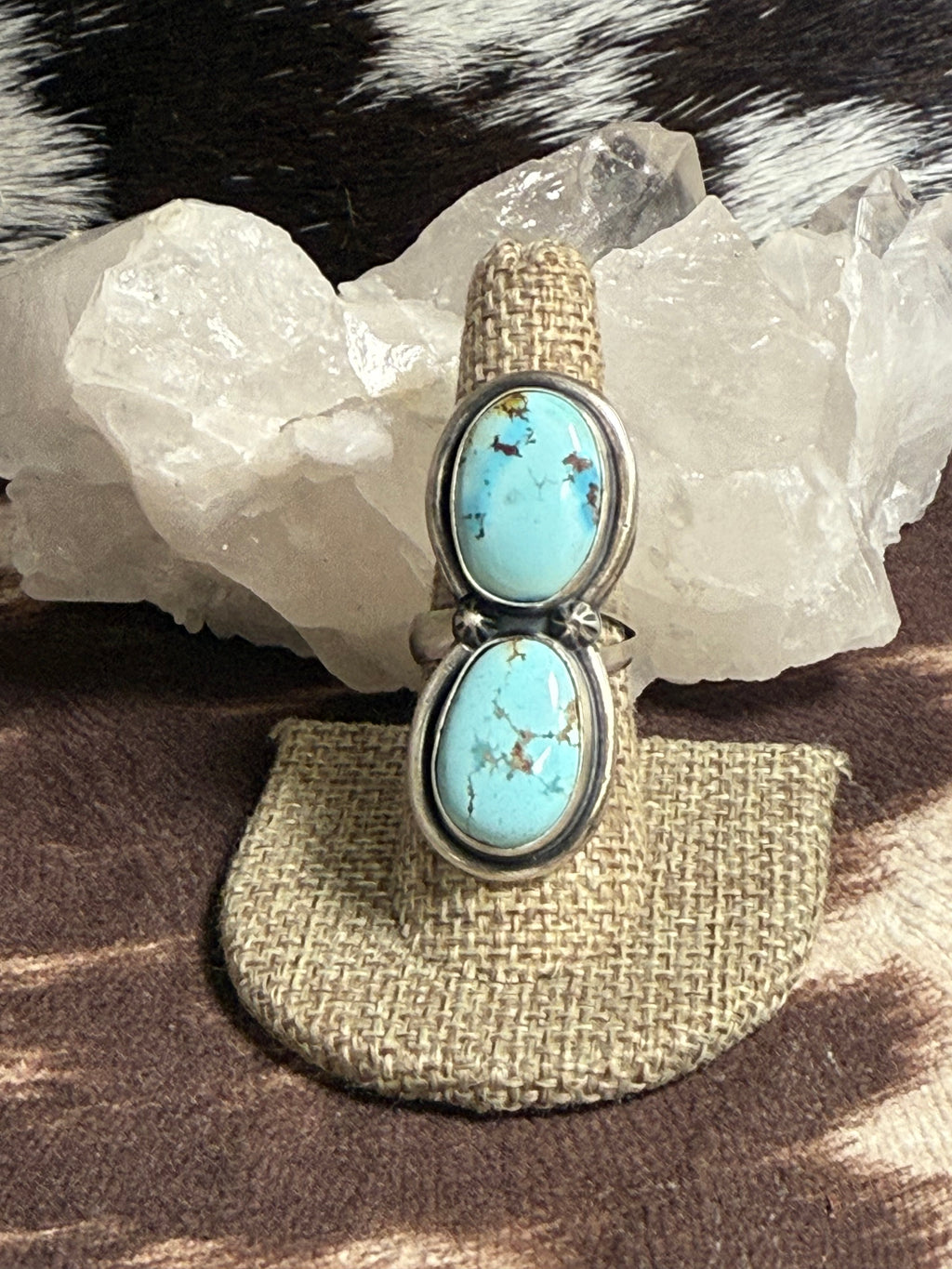 A sterling silver ring with two stacked oval turquoise cabochons, displayed on a burlap ring holder in front of a white crystal cluster.
