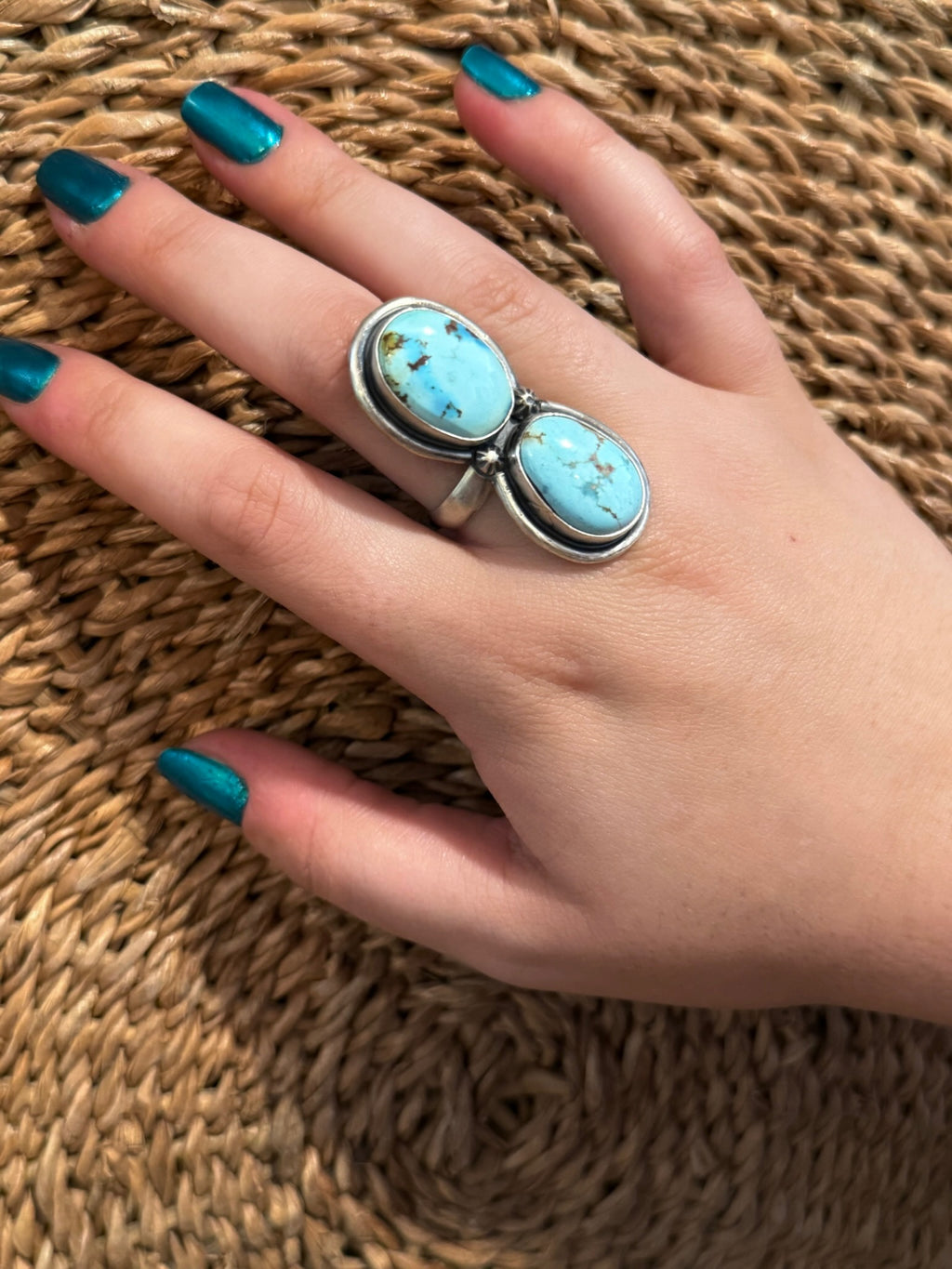 A silver ring with two oval turquoise cabochons worn on a hand with teal nail polish, resting on a woven straw surface.