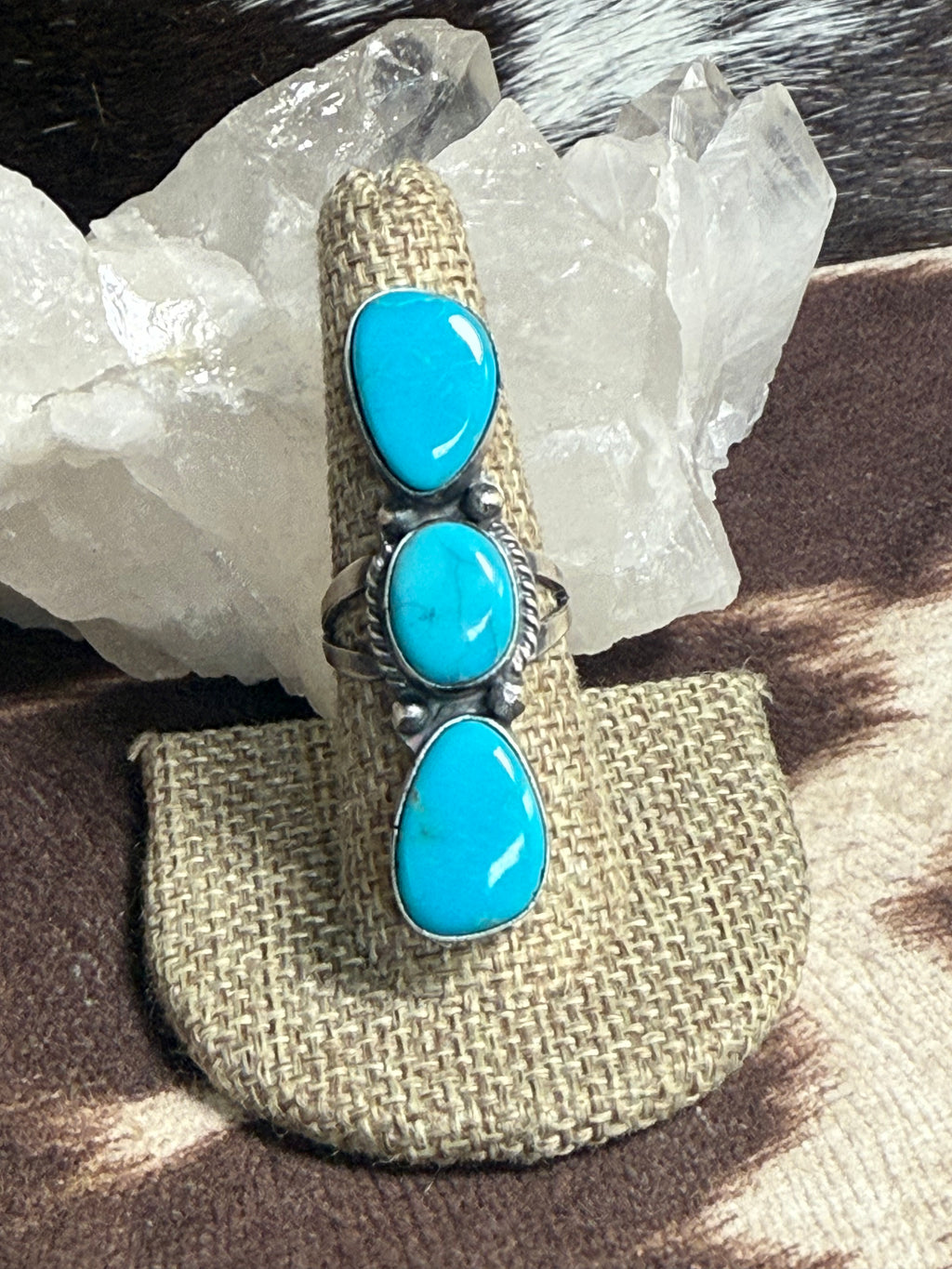 A silver ring with three turquoise stones, displayed on a burlap ring holder. The ring is set against a background of white crystal clusters and a brown and white patterned surface.
