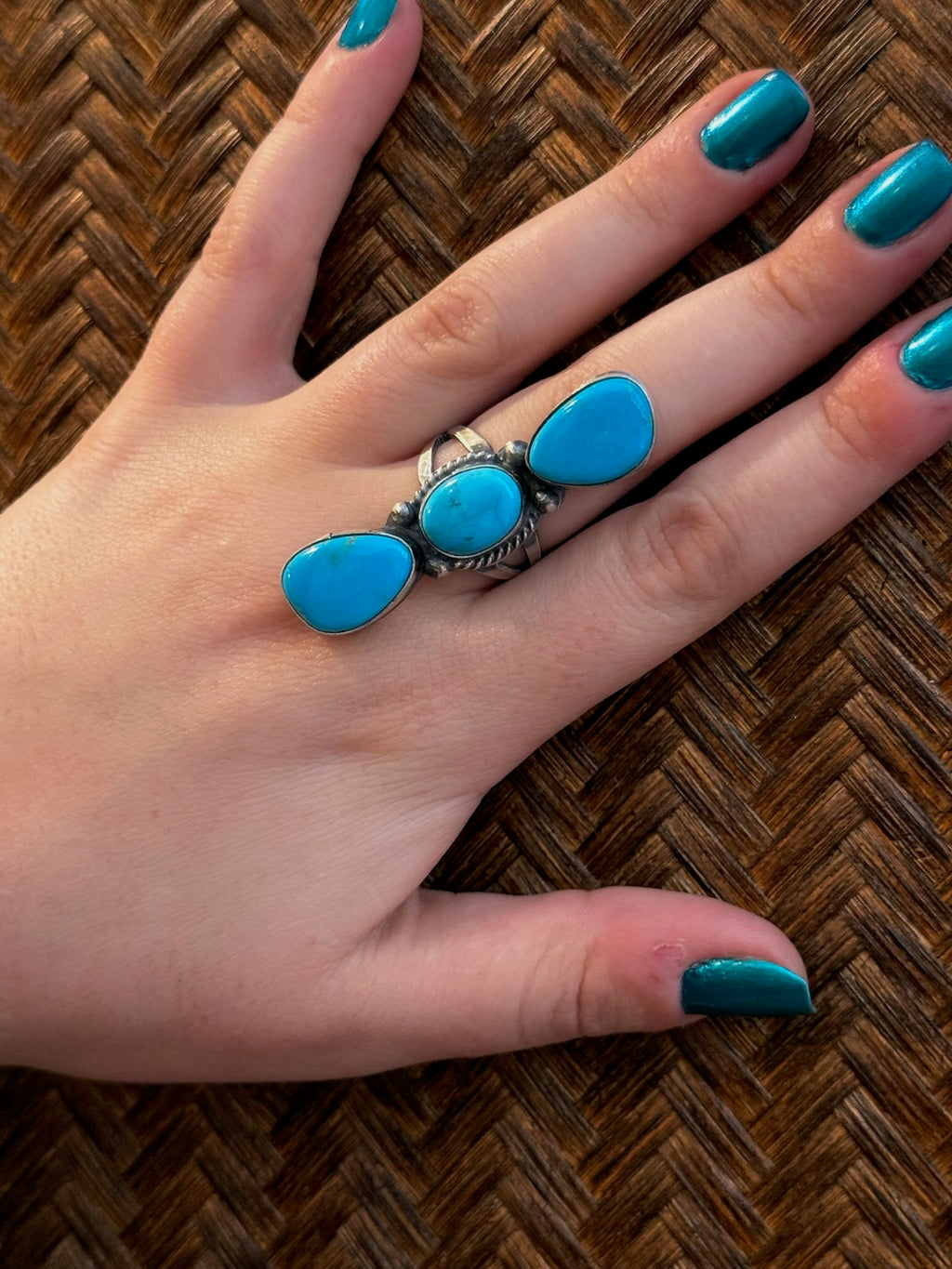 A silver ring with three oval turquoise stones worn on a hand with teal nail polish, resting on a brown woven herringbone surface.