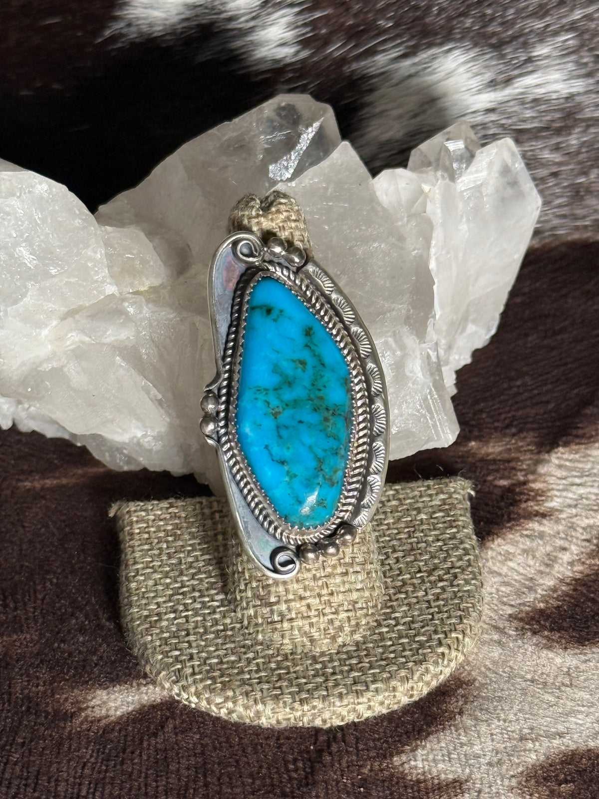 A silver ring featuring a large, teardrop-shaped turquoise stone with natural brown veining, displayed on a textured beige stand in front of white crystal clusters.