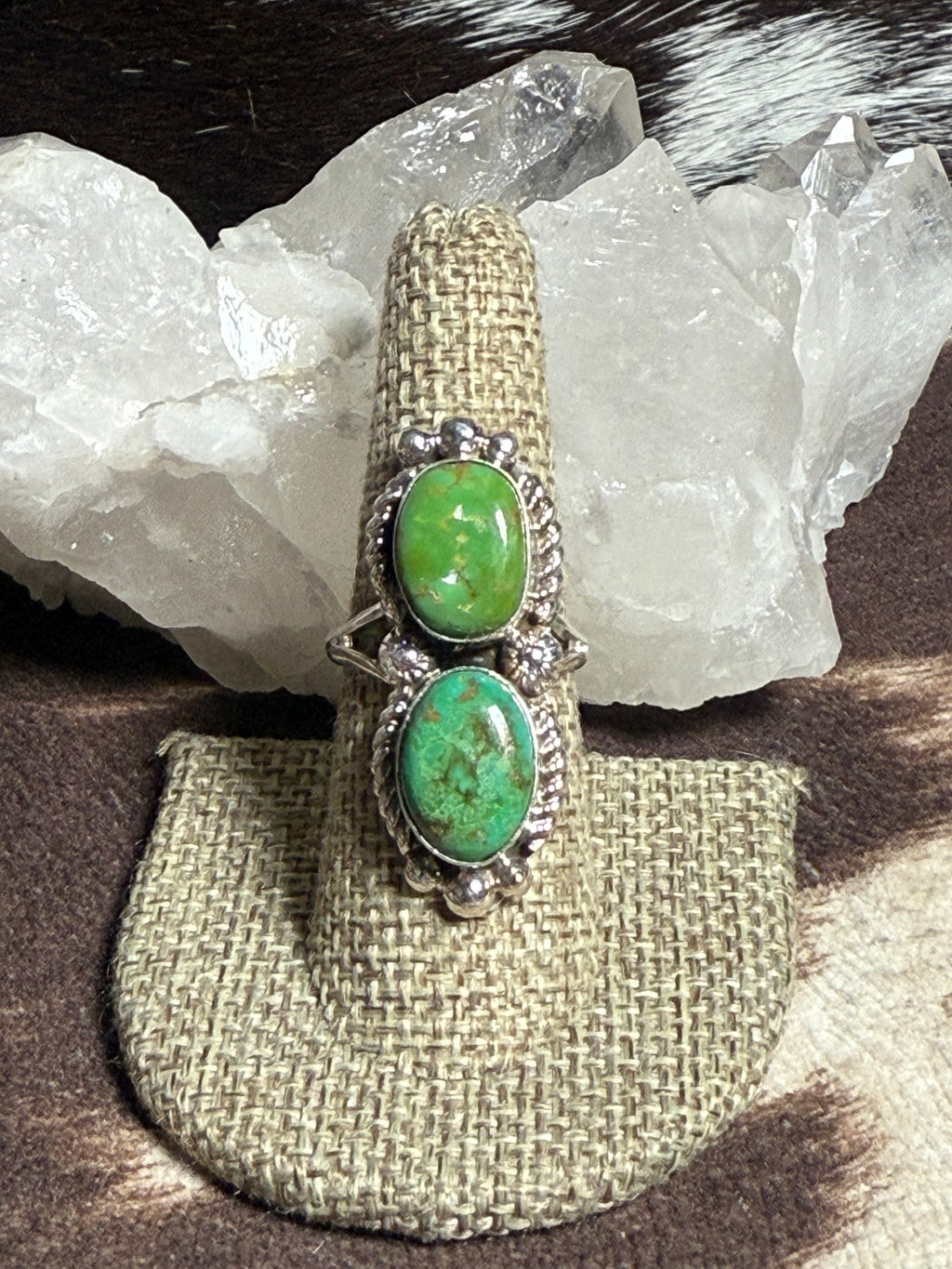 A silver ring with two oval green turquoise stones, displayed on a burlap ring holder in front of white crystal clusters.