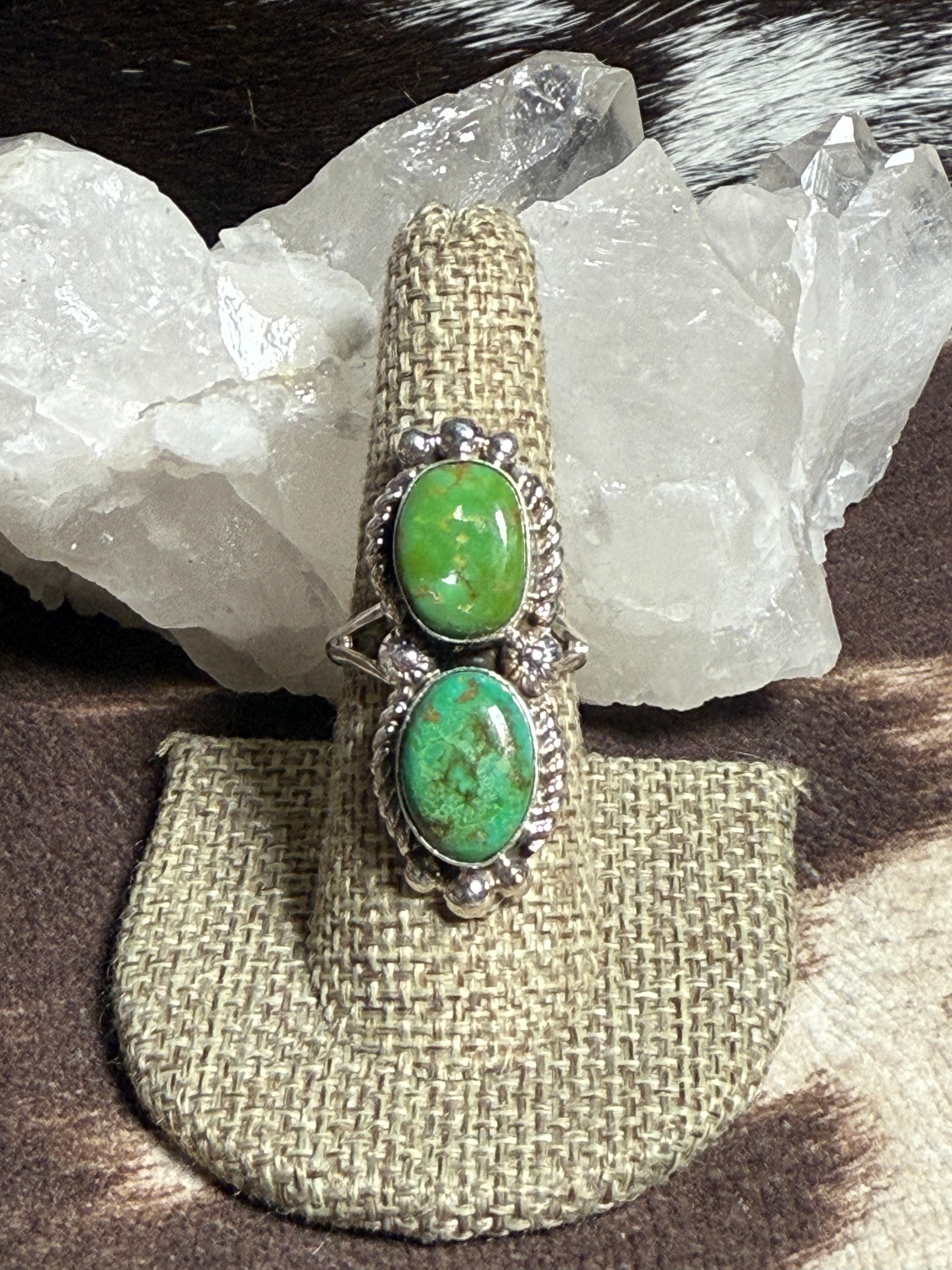 A silver ring with two oval green turquoise stones, displayed on a burlap ring holder in front of white crystal clusters.