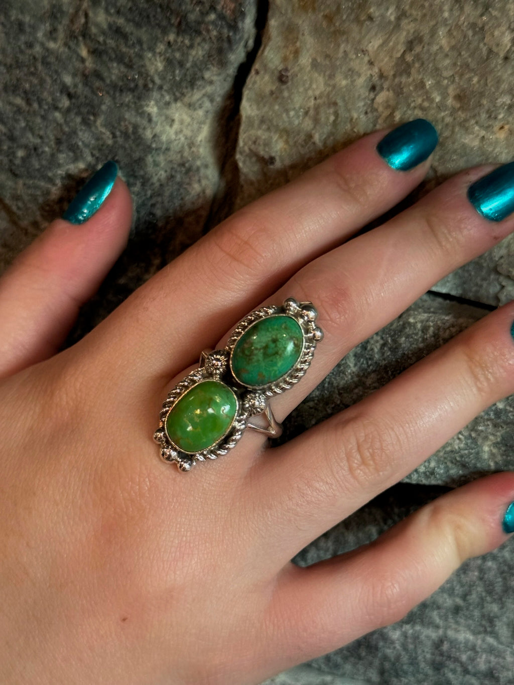 A close-up of a hand with metallic teal nail polish wearing a silver ring with two oval green cabochon gemstones, set against a textured gray stone background.