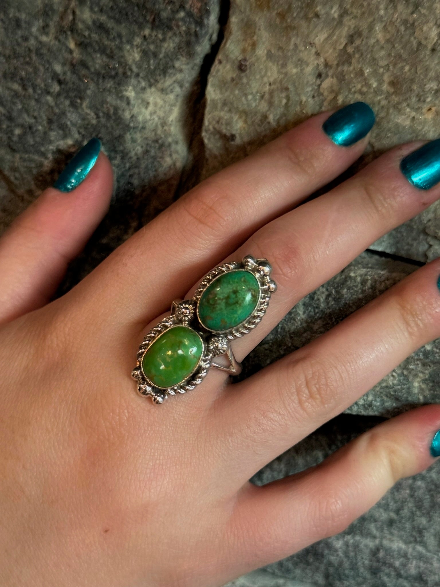 A close-up of a hand with metallic teal nail polish wearing a silver ring with two oval green cabochon gemstones, set against a textured gray stone background.