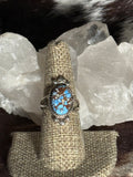 A silver ring with an oval turquoise and brown stone center, displayed on a burlap ring holder in front of white crystal clusters.