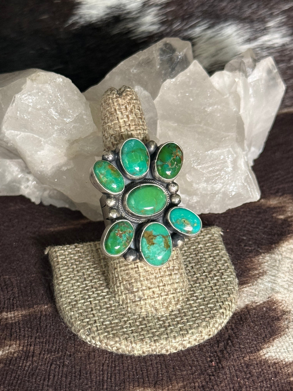 A silver ring with seven green turquoise stones, displayed on a burlap ring holder in front of white crystal clusters.