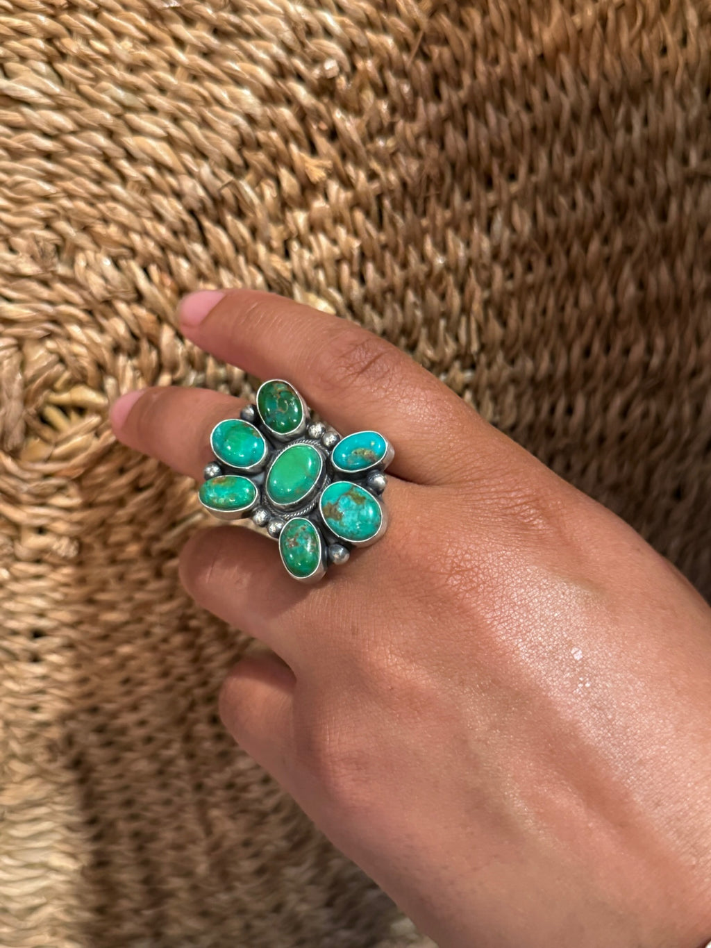 A close-up of a hand wearing a large silver ring with seven bright turquoise-colored stones, set against a woven straw background.