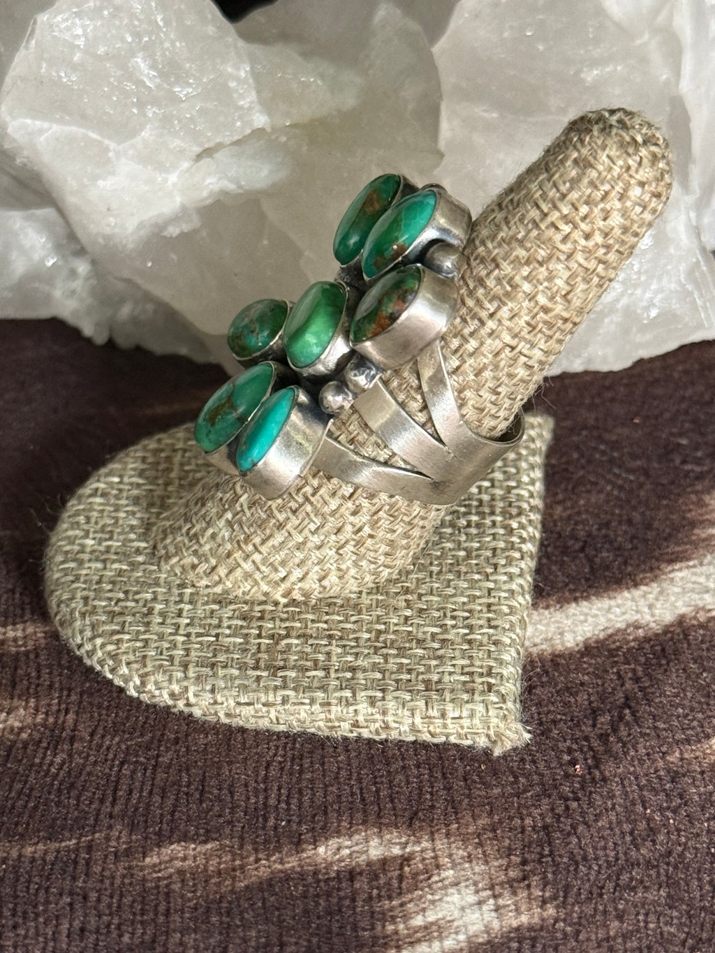 A silver ring with multiple oval green turquoise-like stones, displayed on a woven beige ring stand over a brown textured surface with white crystal formations in the background.