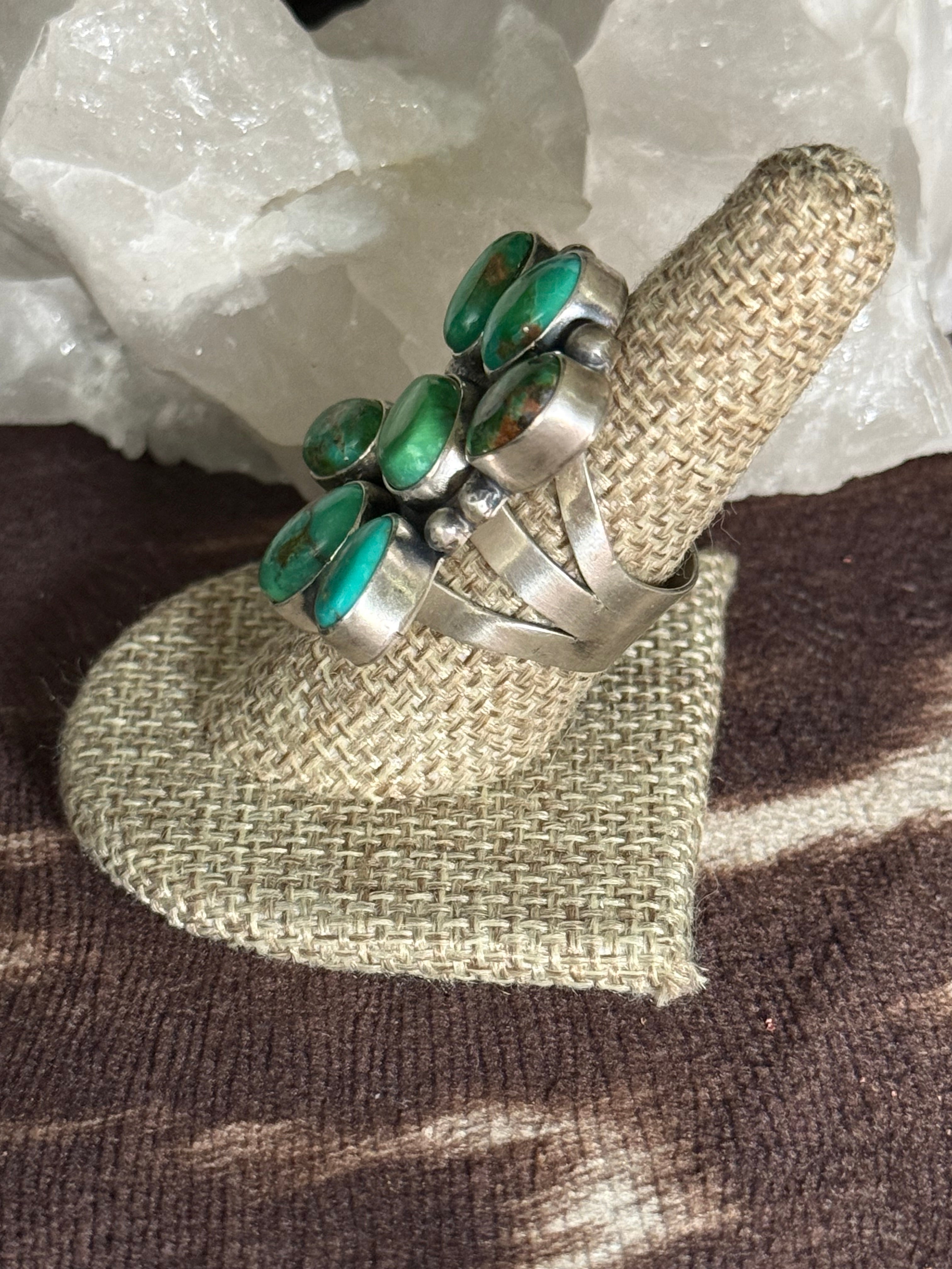 A silver ring with multiple oval green turquoise-like stones, displayed on a woven beige ring stand over a brown textured surface with white crystal formations in the background.