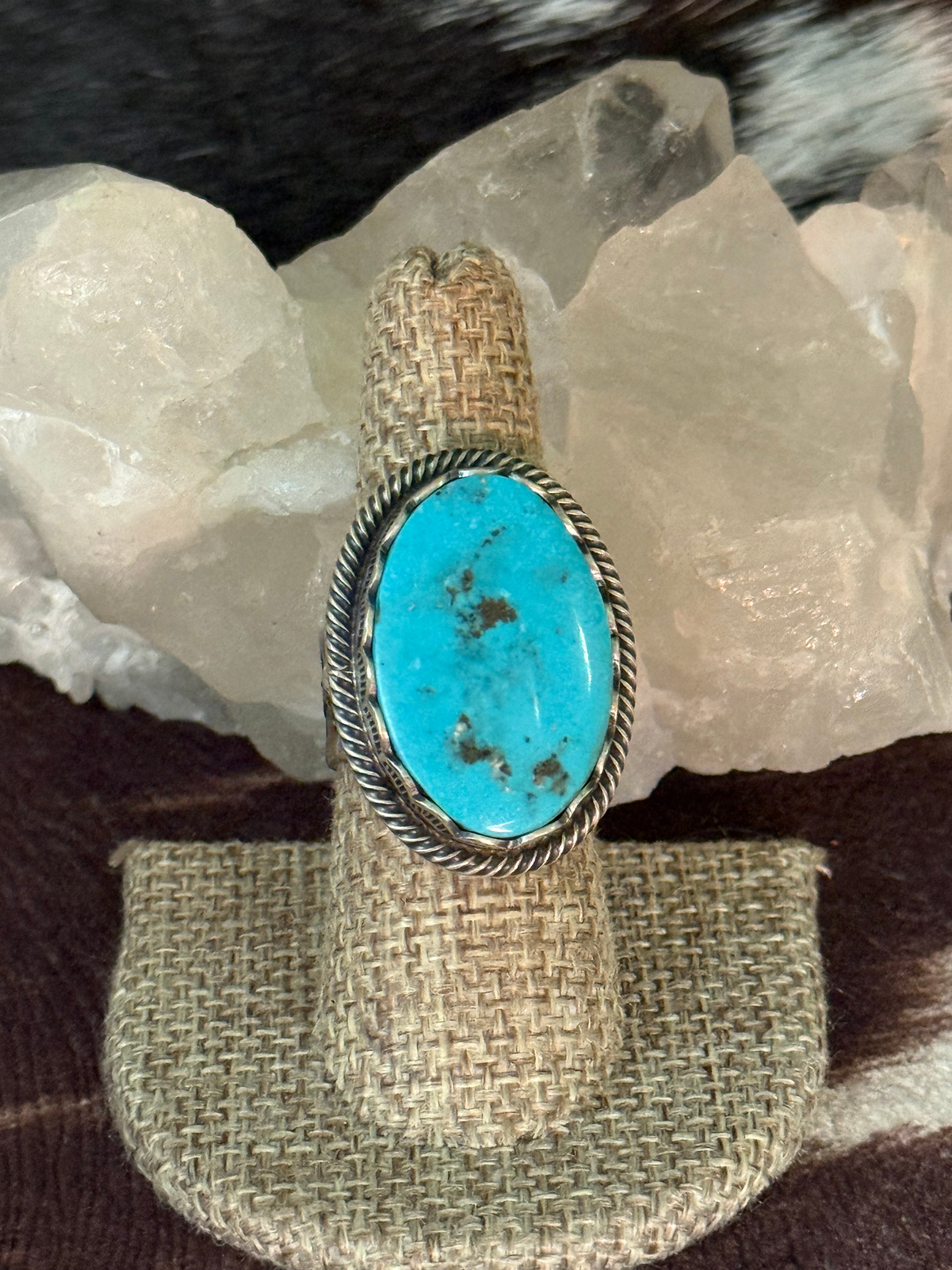 A silver ring with a large oval turquoise stone, displayed on a woven ring stand in front of white crystal rocks.