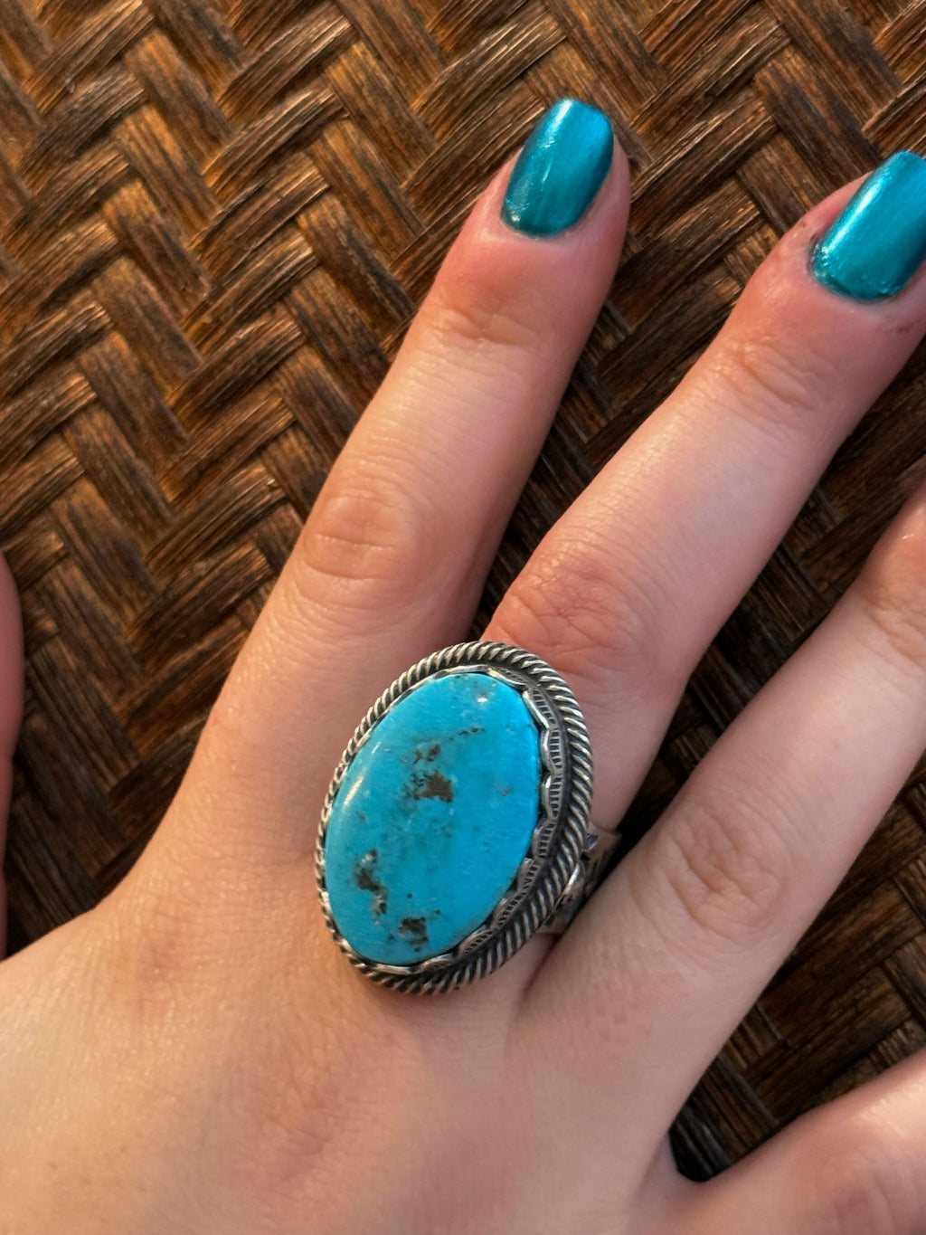 A close-up of a hand wearing a silver ring with a large oval turquoise stone, resting on a brown woven surface.