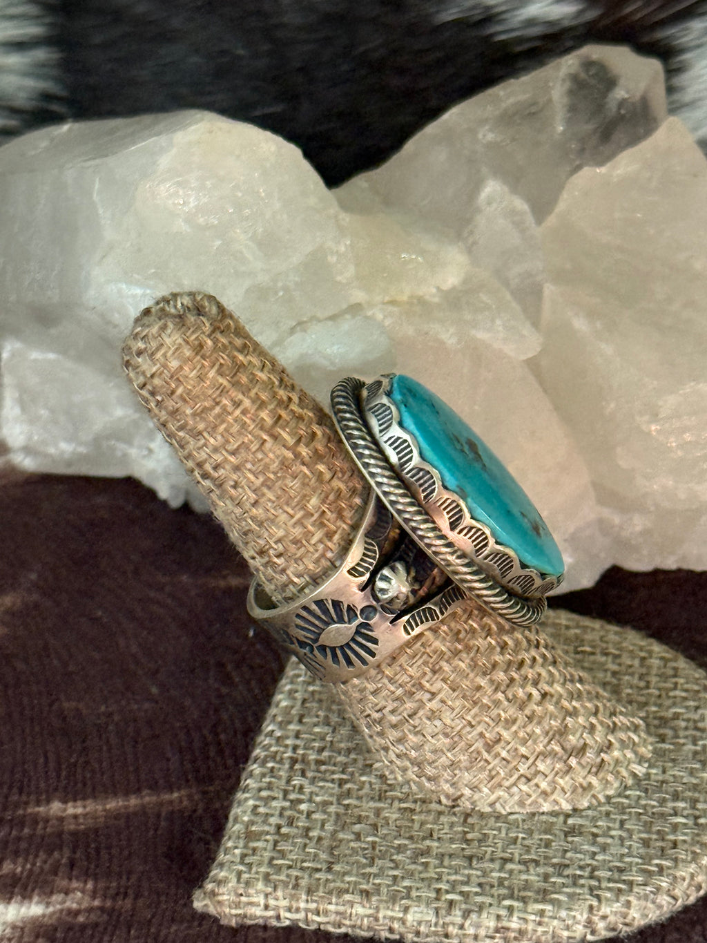A silver ring with a turquoise stone and engraved details, displayed on a burlap ring holder with white crystal rocks in the background.