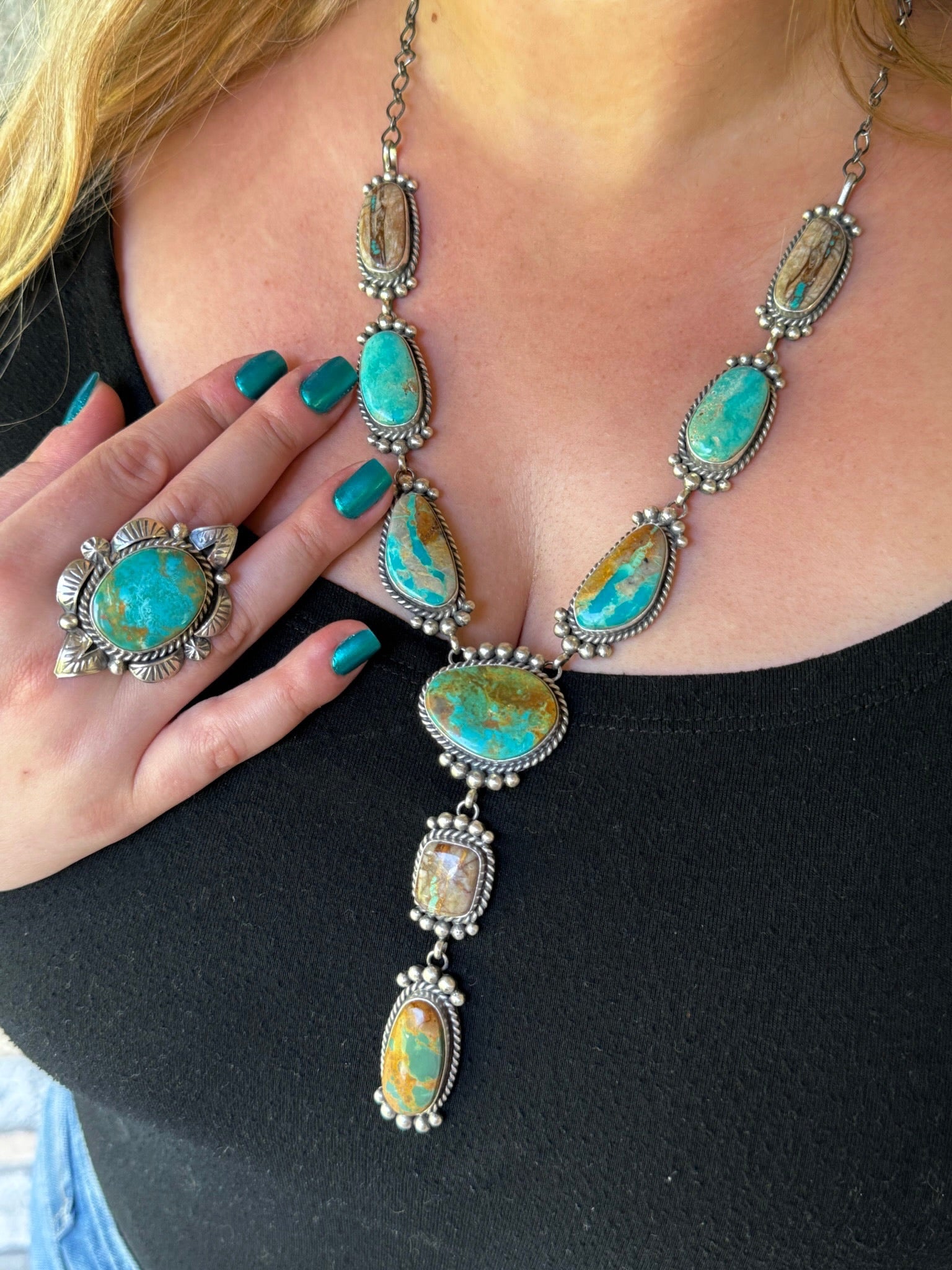 A close-up of a person wearing a silver-toned statement necklace and matching ring, both featuring large turquoise-colored stone clusters with natural brown veining. The person is wearing a black top, and the jewelry is displayed prominently against the neck and hand.